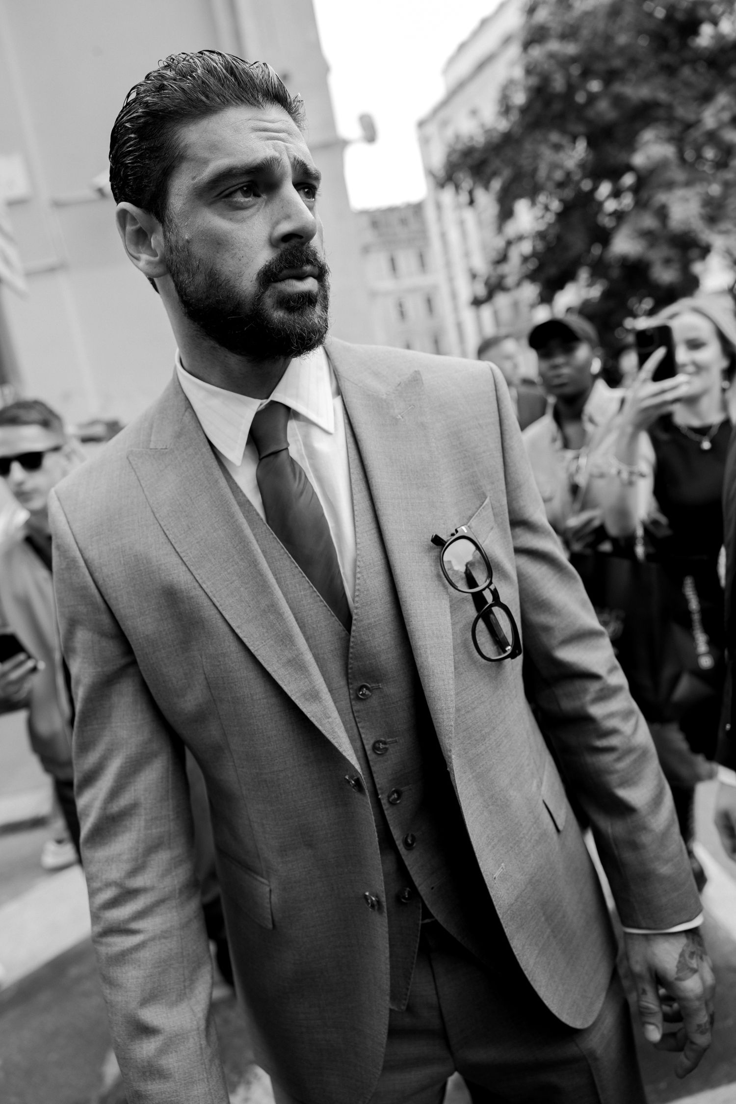 Well-dressed man in three piece gray suit walking outdoors in black and white photograph with sunglasses tucked in pocket.