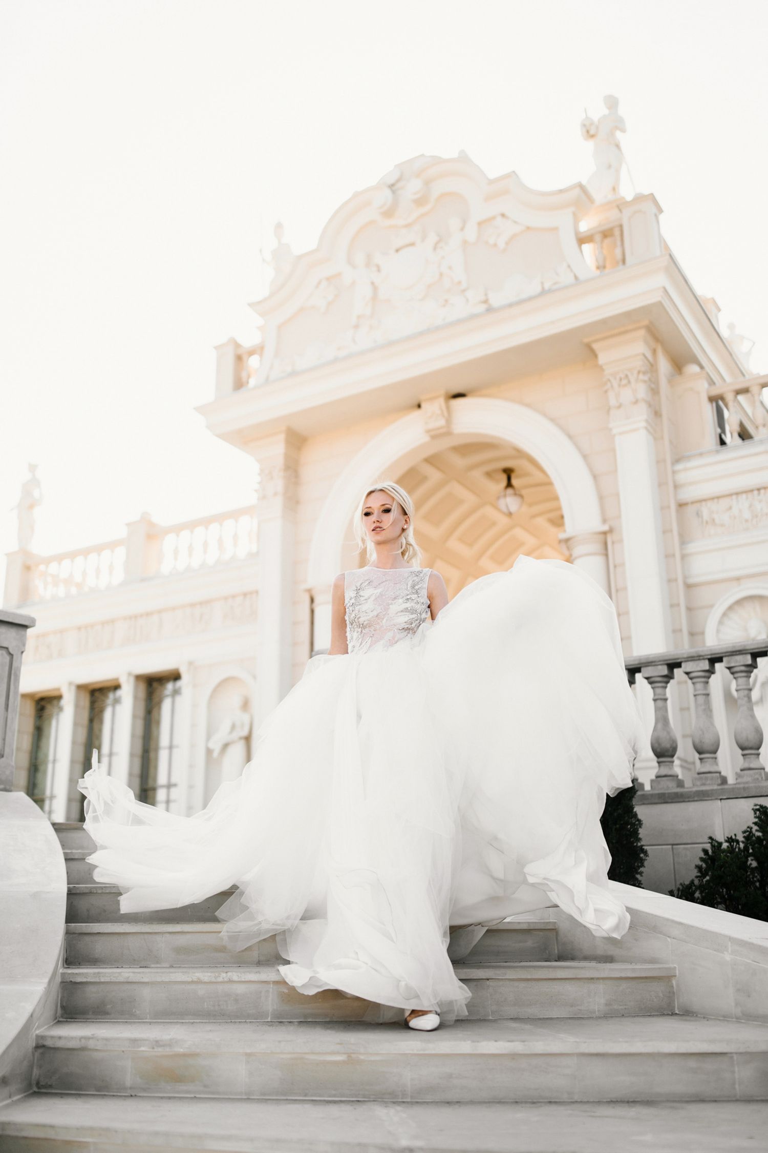 Flowing white wedding dress captured on ornate white marble staircase with architectural details.