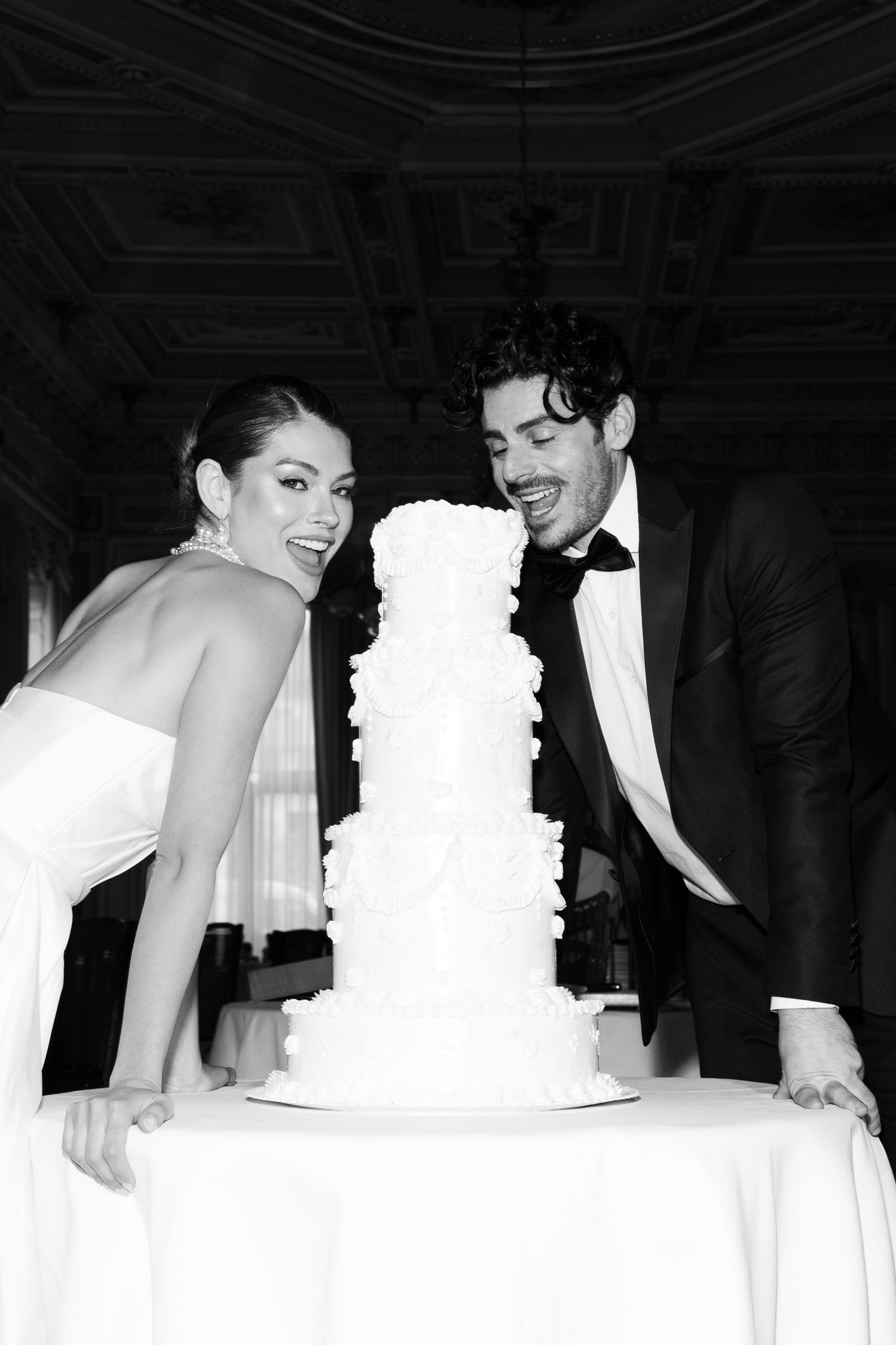 Black and white photo of a newlywed couple playfully posing with their tall white tiered wedding cake.