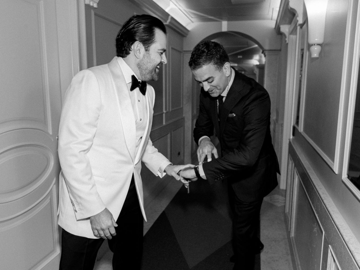 Two men in formal wear checking time in an elegant hallway.
