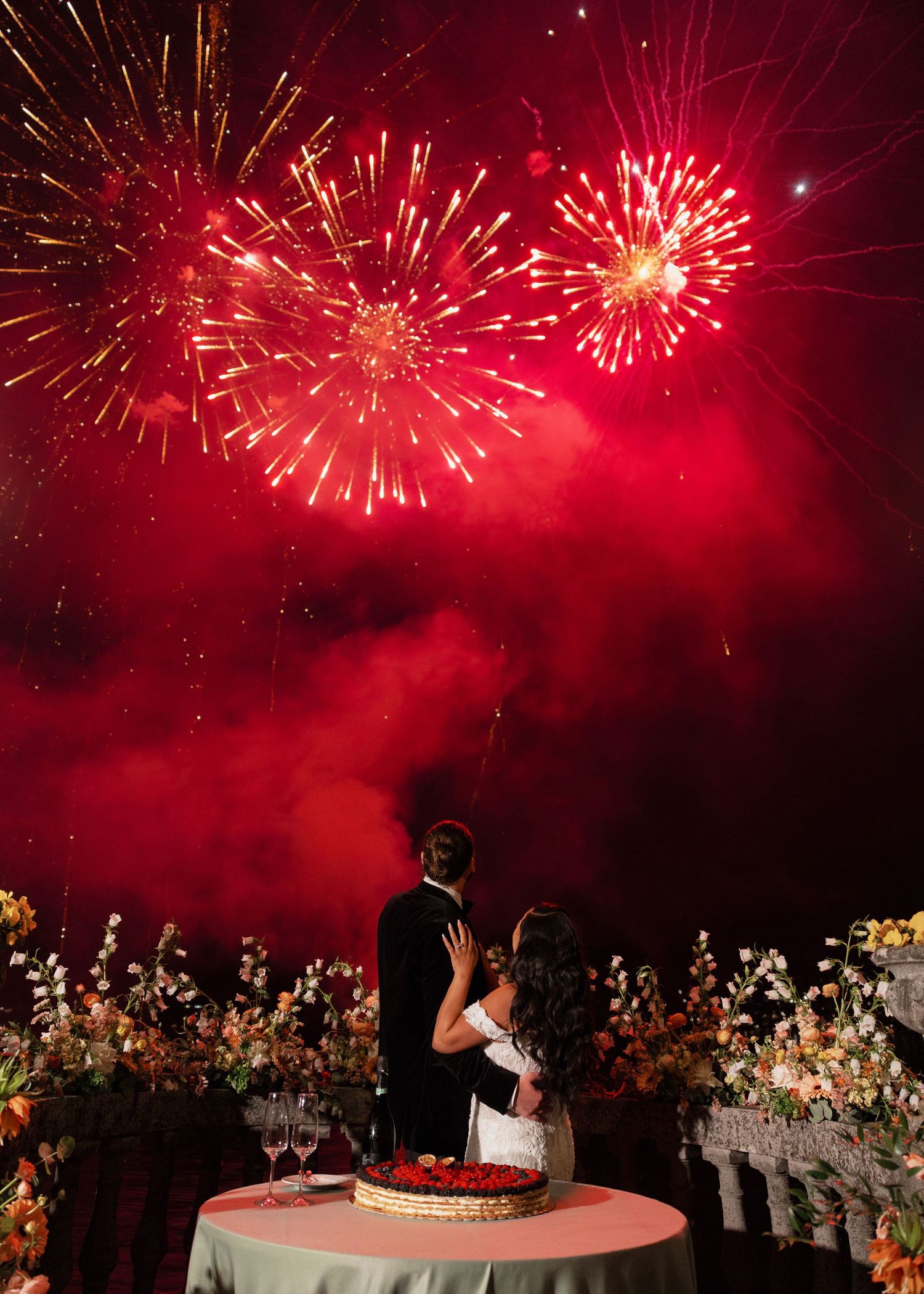 Bride and groom watching stunning red fireworks display at their wedding reception with floral arrangements.