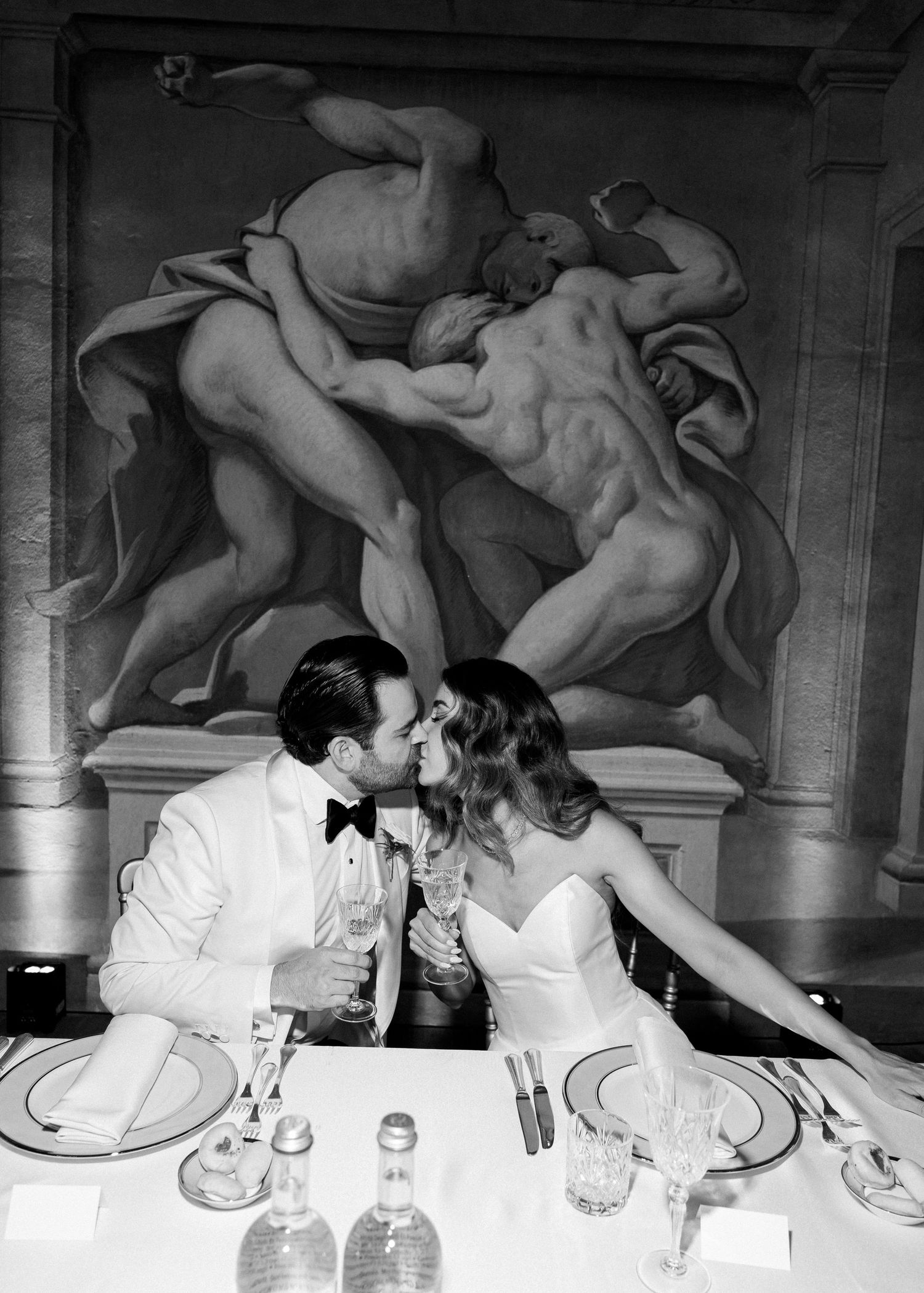 Black and white photo of a couple kissing at a wedding reception table beneath a classical Renaissance mural.