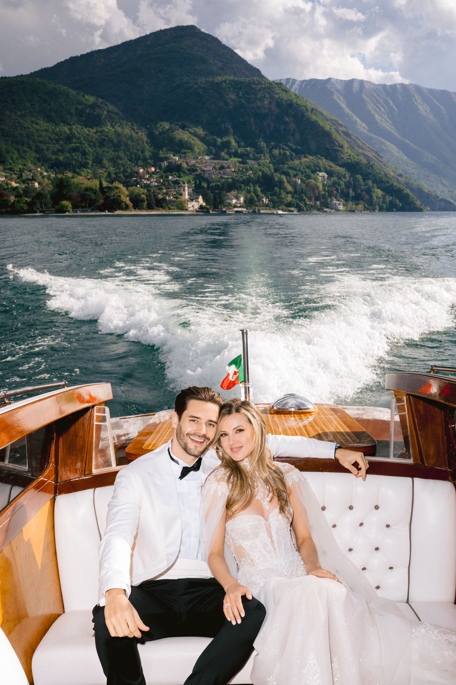 Bride and groom sitting on a luxury wooden boat on Lake Como, Italy, with mountain backdrop.