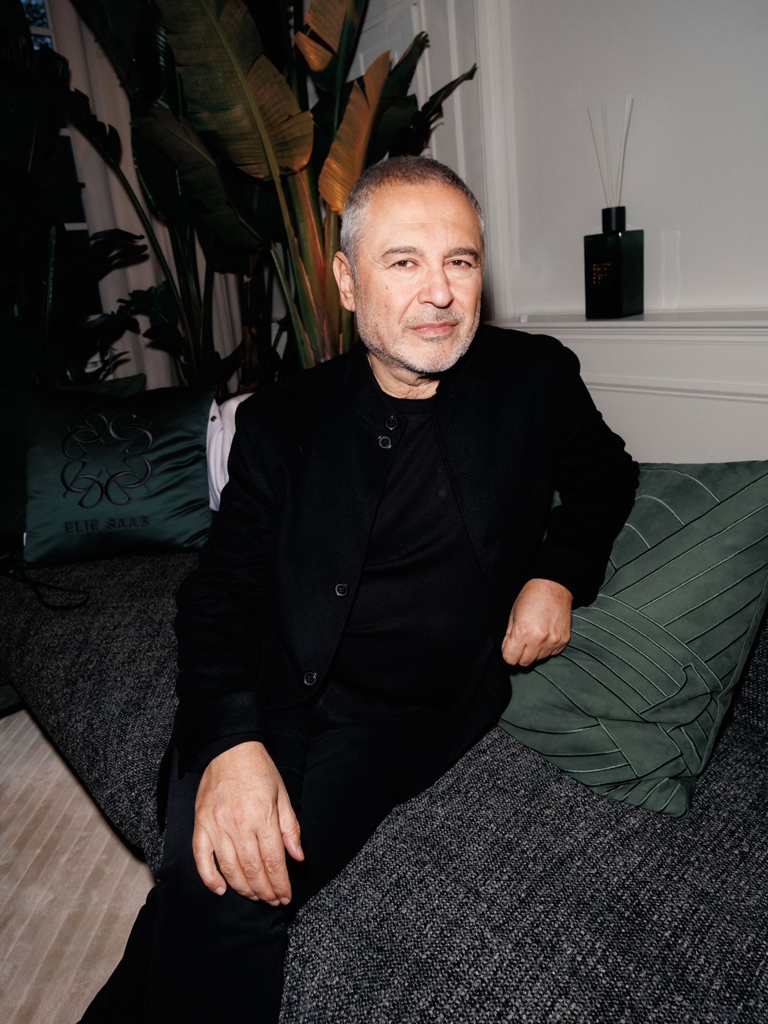 A person wearing black clothing sits casually on a dark gray couch with green pillows in a modern interior setting.