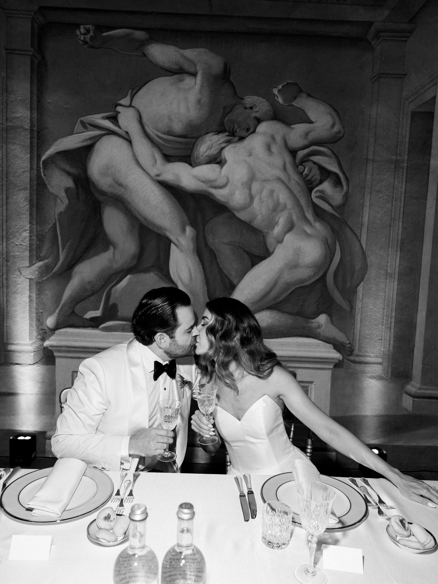 Romantic black and white photo of a couple dining beneath classical artwork in formal evening attire.