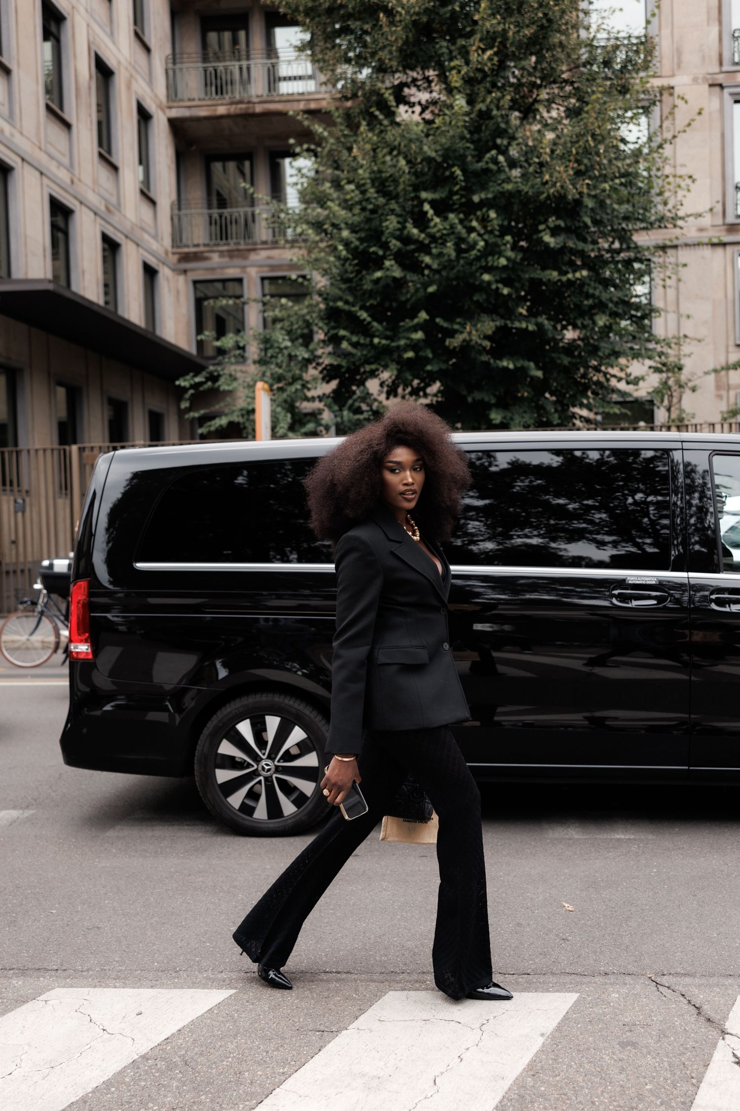A woman in a black business suit strikes a pose on a crosswalk with a luxury van in the background on a city street.