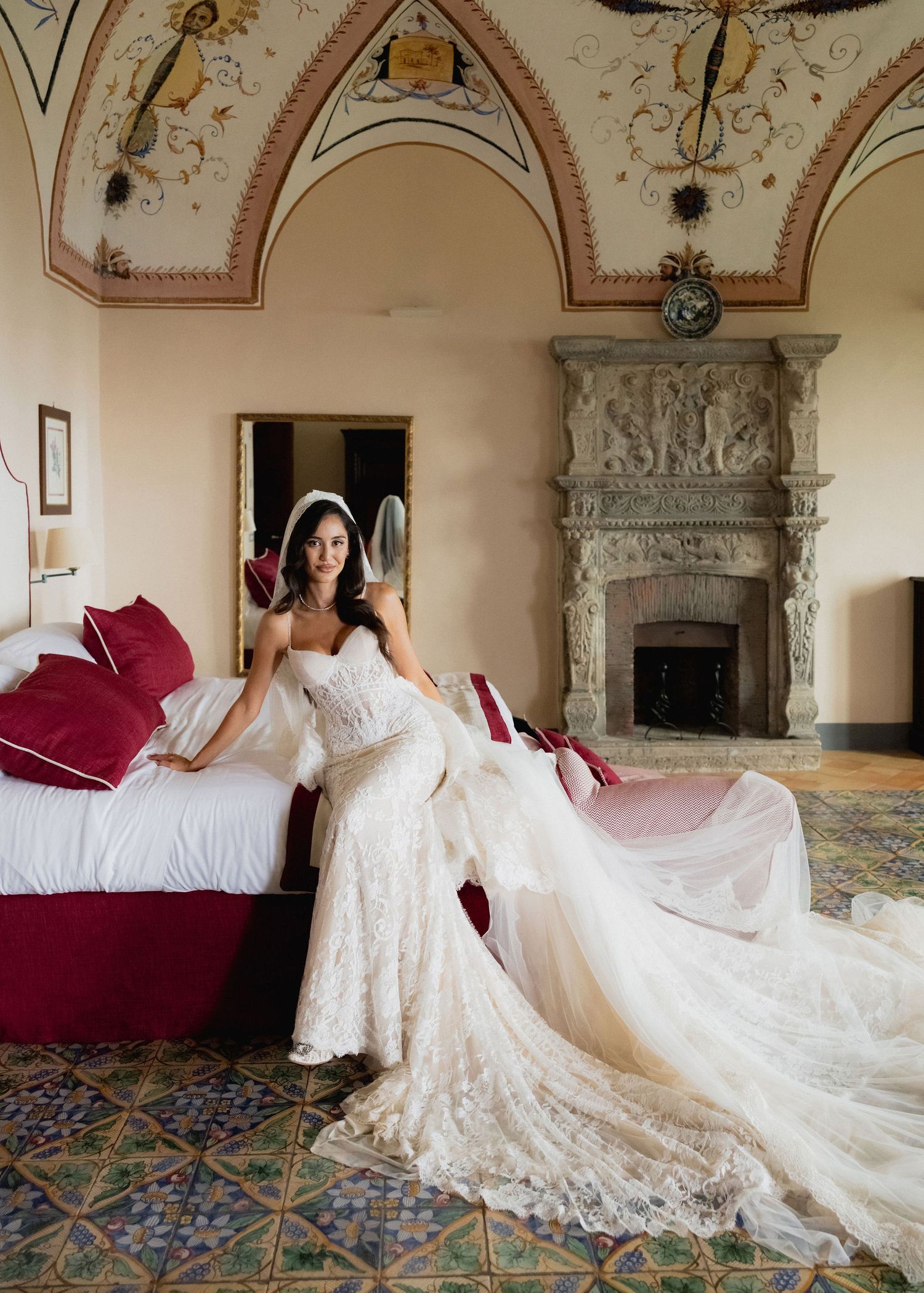 Bride in lace wedding gown with long train sits beside ornate fireplace in elegant Italian villa room.