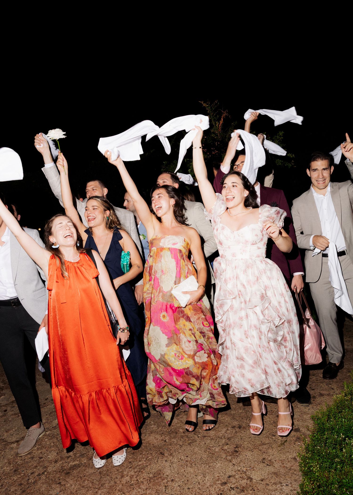 Joyful wedding guests in colorful dresses wave white napkins in the air during an outdoor evening celebration.