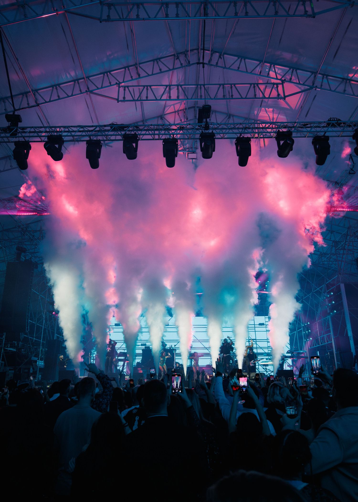 Concert crowd silhouetted against dramatic pink and blue smoke effects with stage lighting overhead.