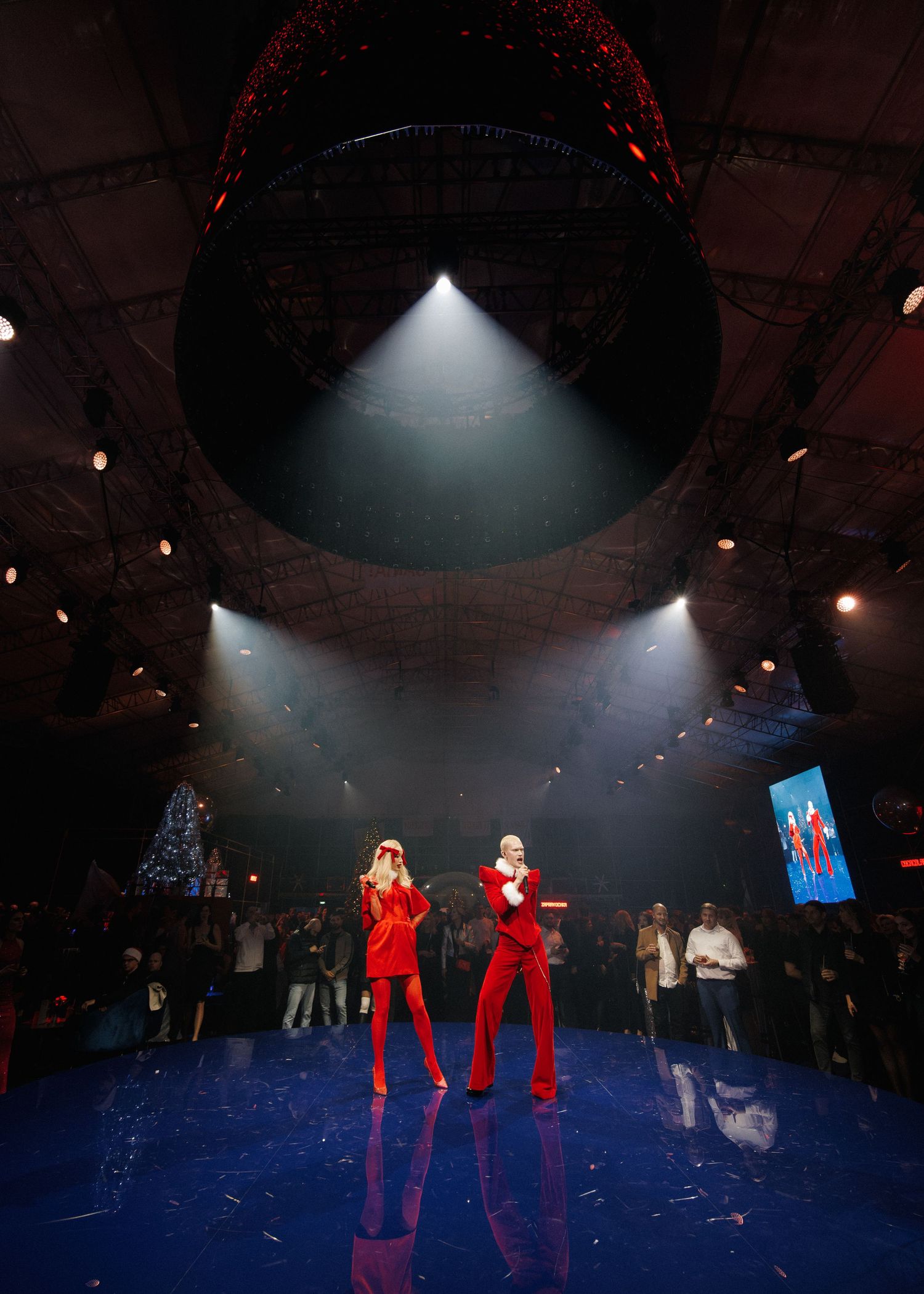 Two performers in red outfits on a dark stage with dramatic spotlighting and a blue floor.