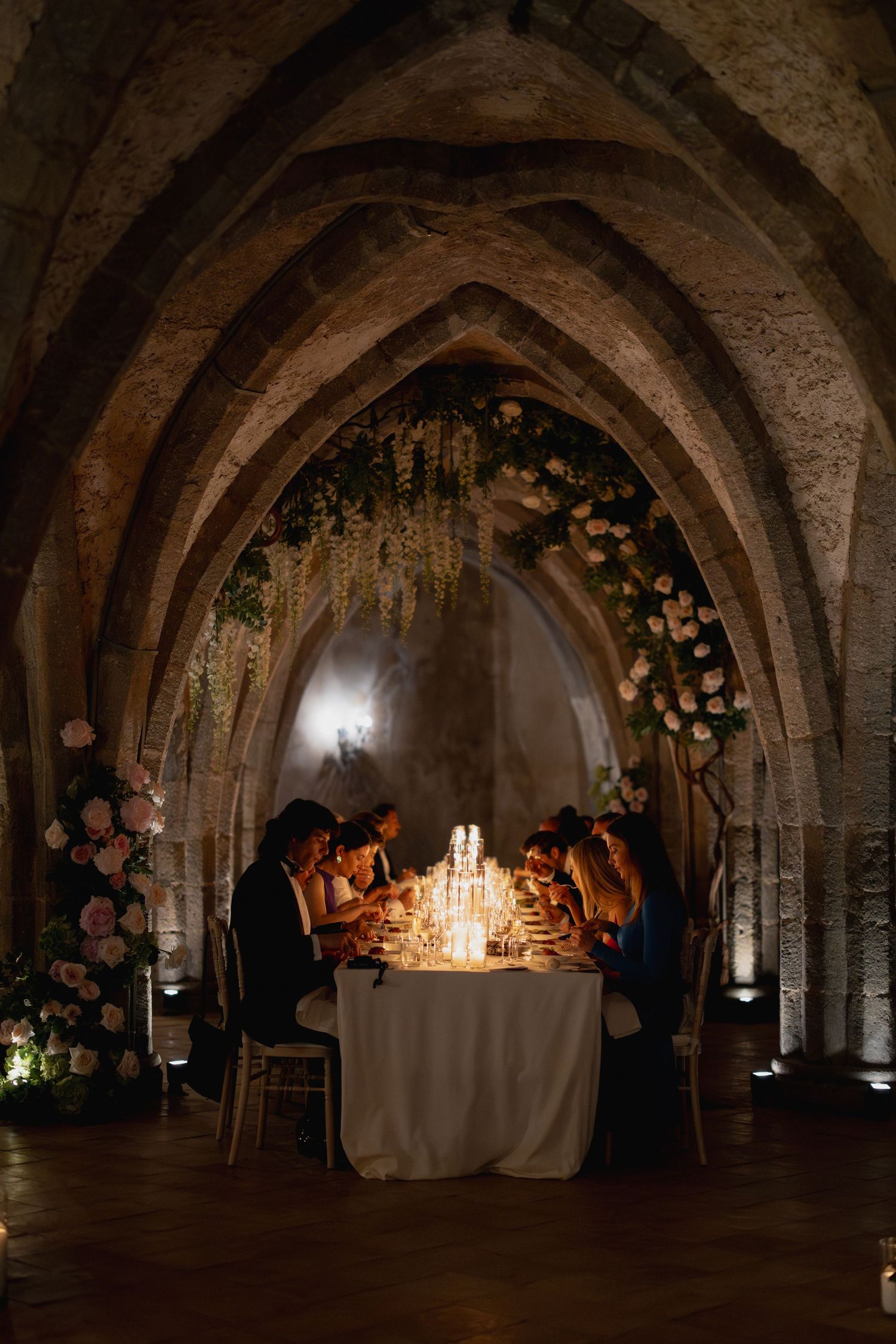 Candlelit medieval stone crypt with arched ceilings decorated with flowers and tables set for an intimate dinner gathering.