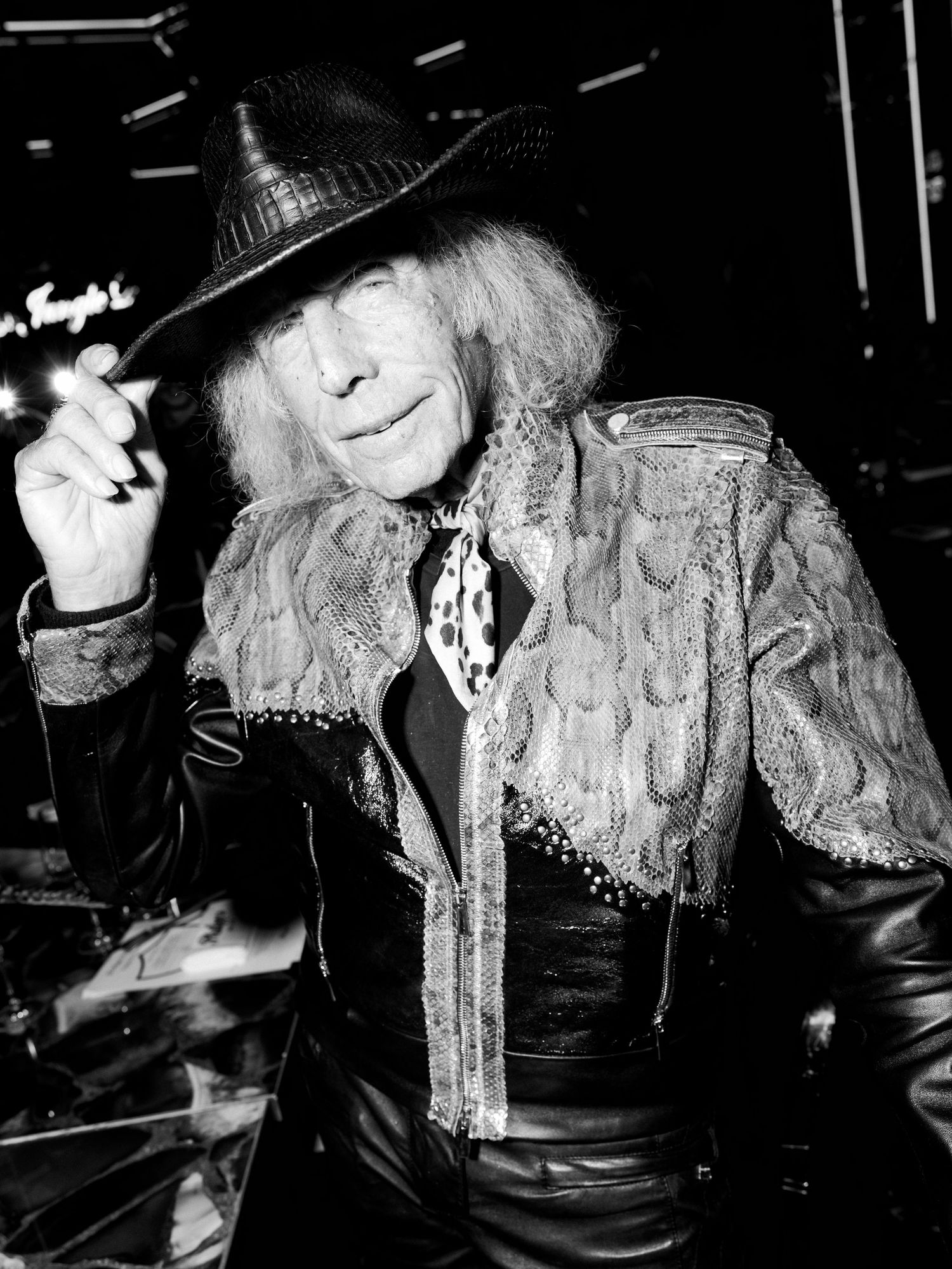 A black and white photo of someone in a leather jacket and hat smoking a cigarette at a bar.