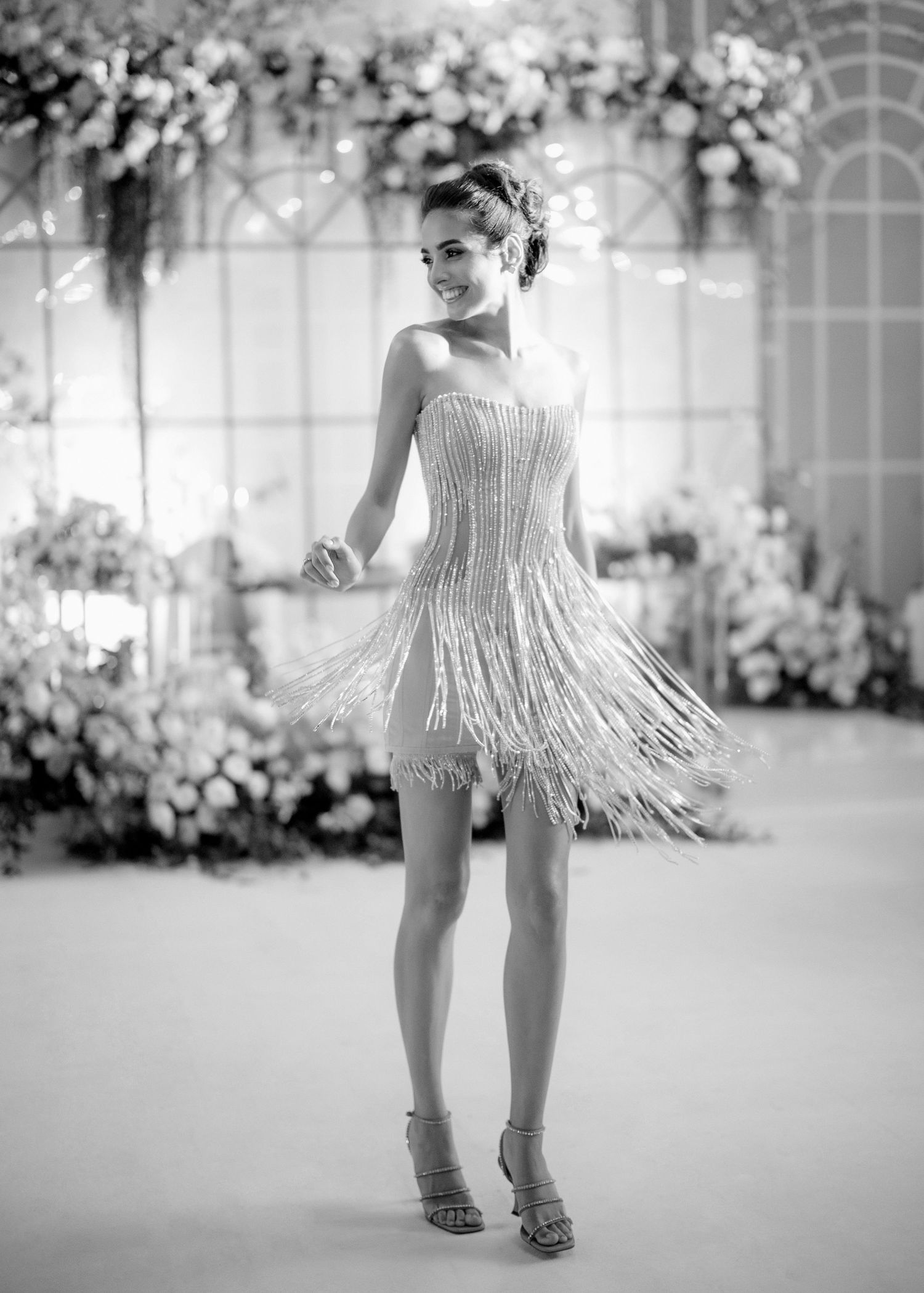 Woman in white beaded fringe mini dress posing at elegant floral event in black and white photo.