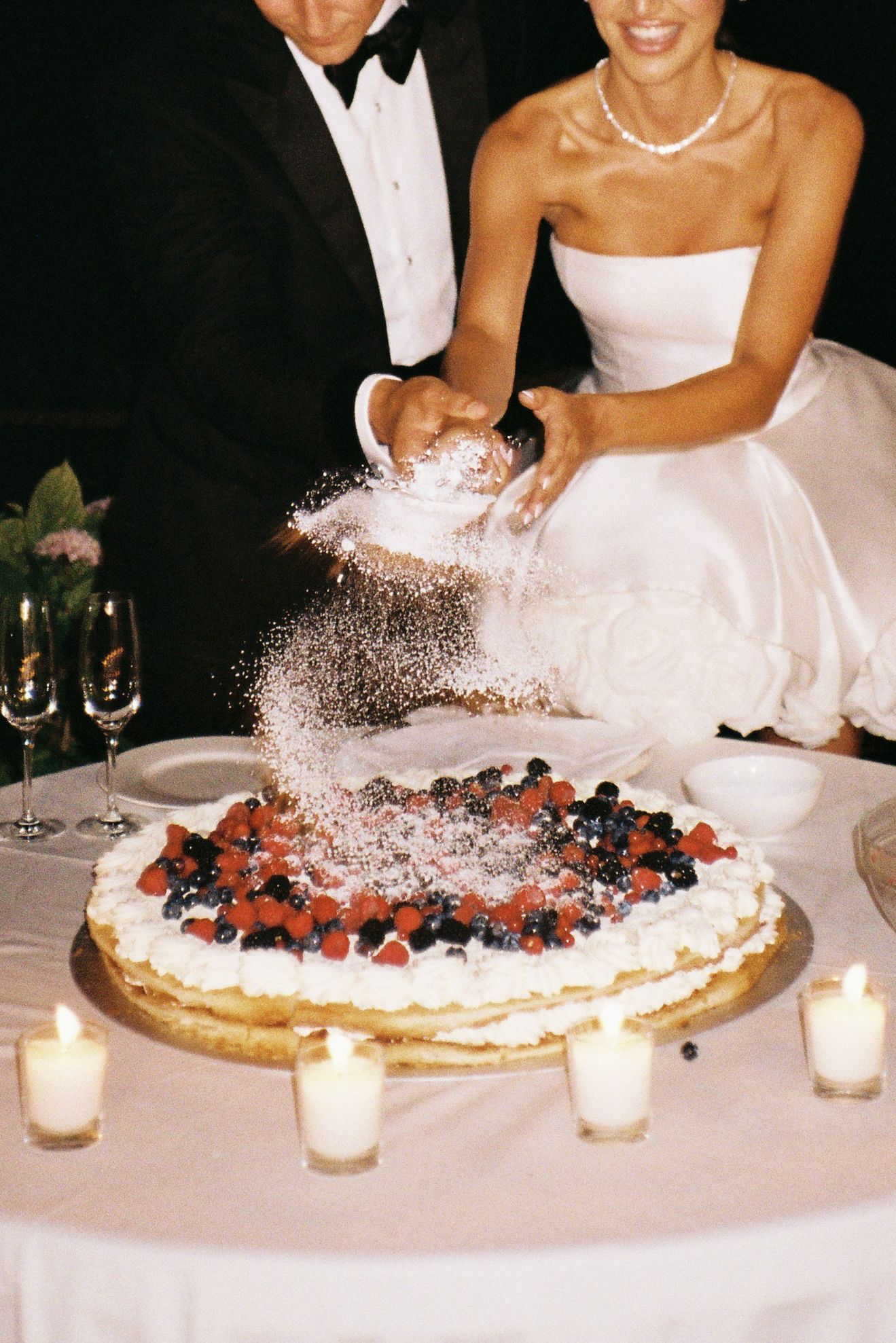 Powdered sugar being sprinkled over a berry-topped wedding cake surrounded by candlelight.