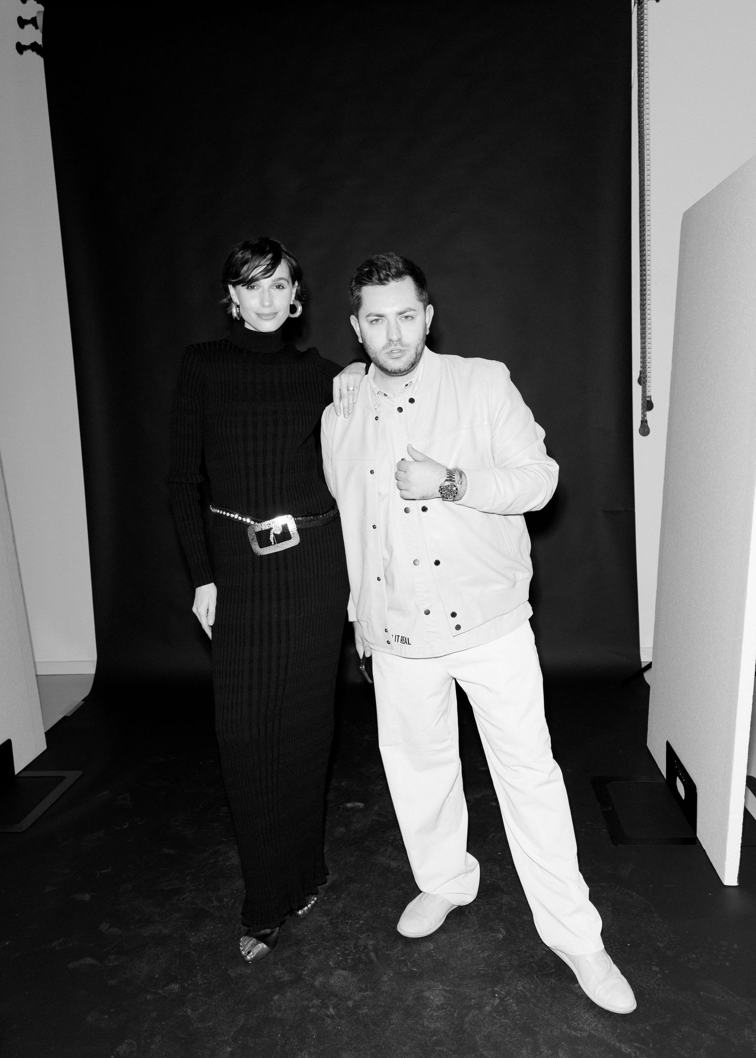 Two people pose against a black backdrop in a black and white photo with contrasting outfits.