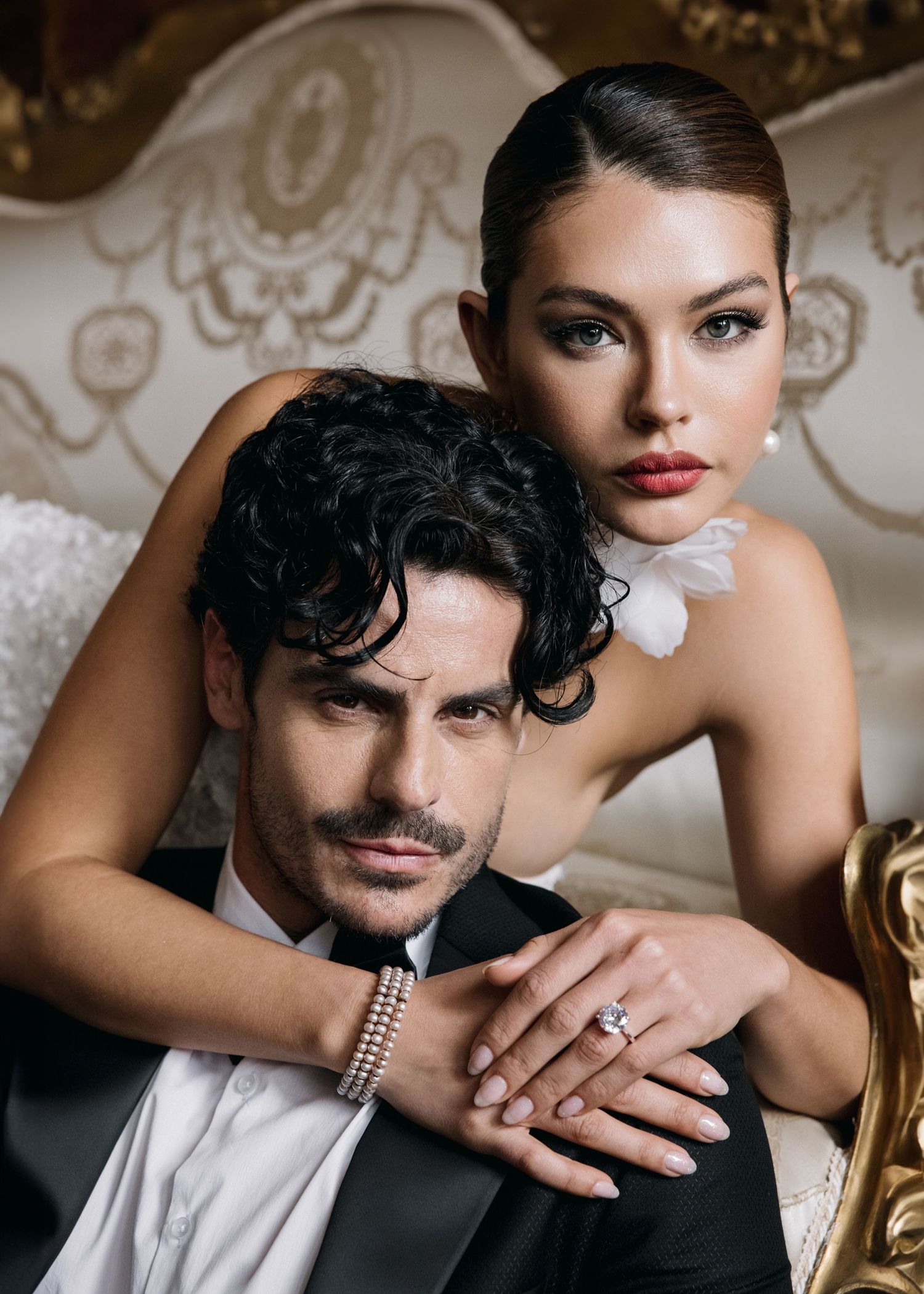 Elegant couple posing in luxury setting, woman in strapless gown with diamond ring, man in black tuxedo with pearl bracelet.