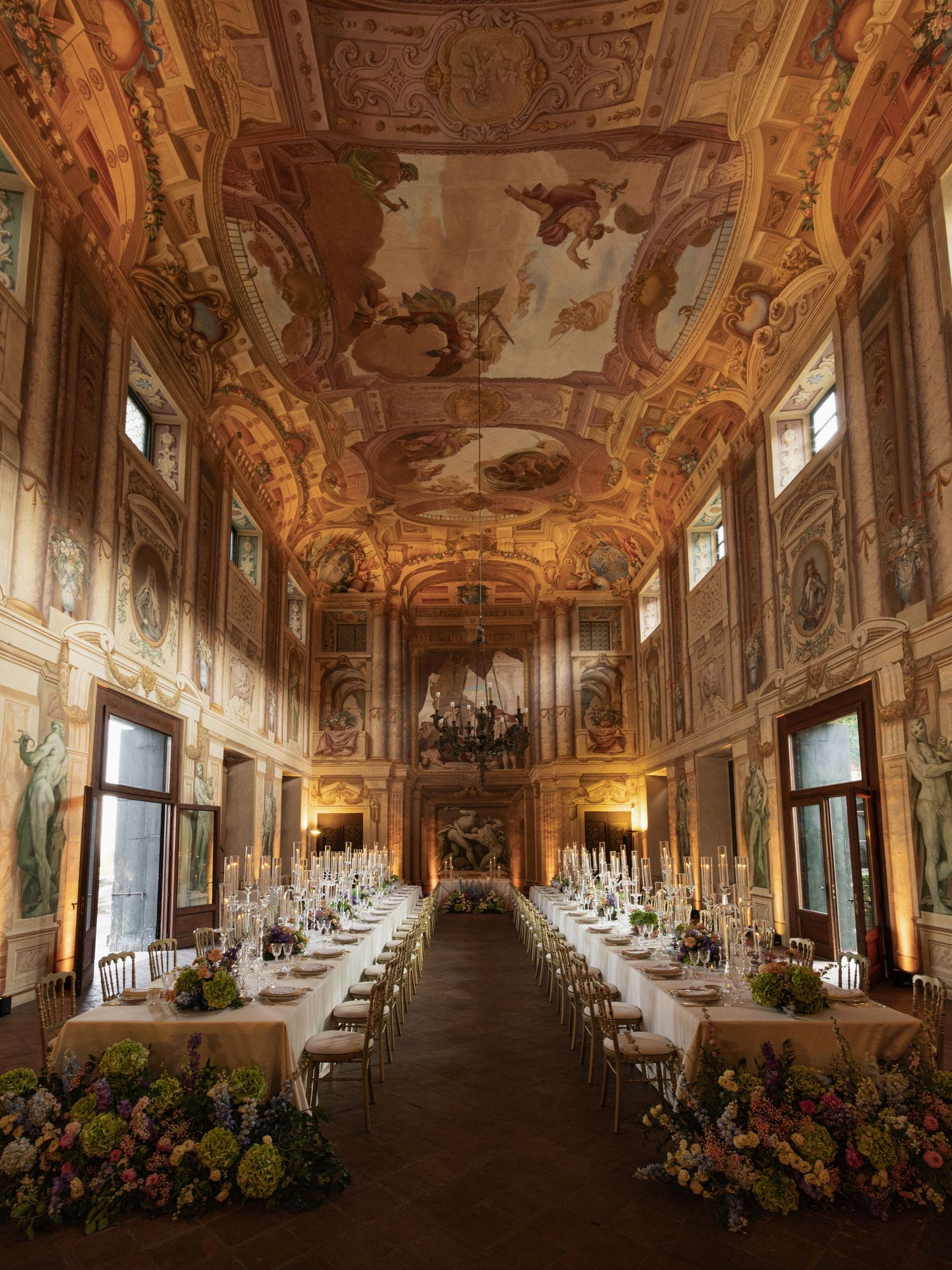Ornate baroque banquet hall with frescoed ceiling, long dining tables adorned with candles and floral arrangements.