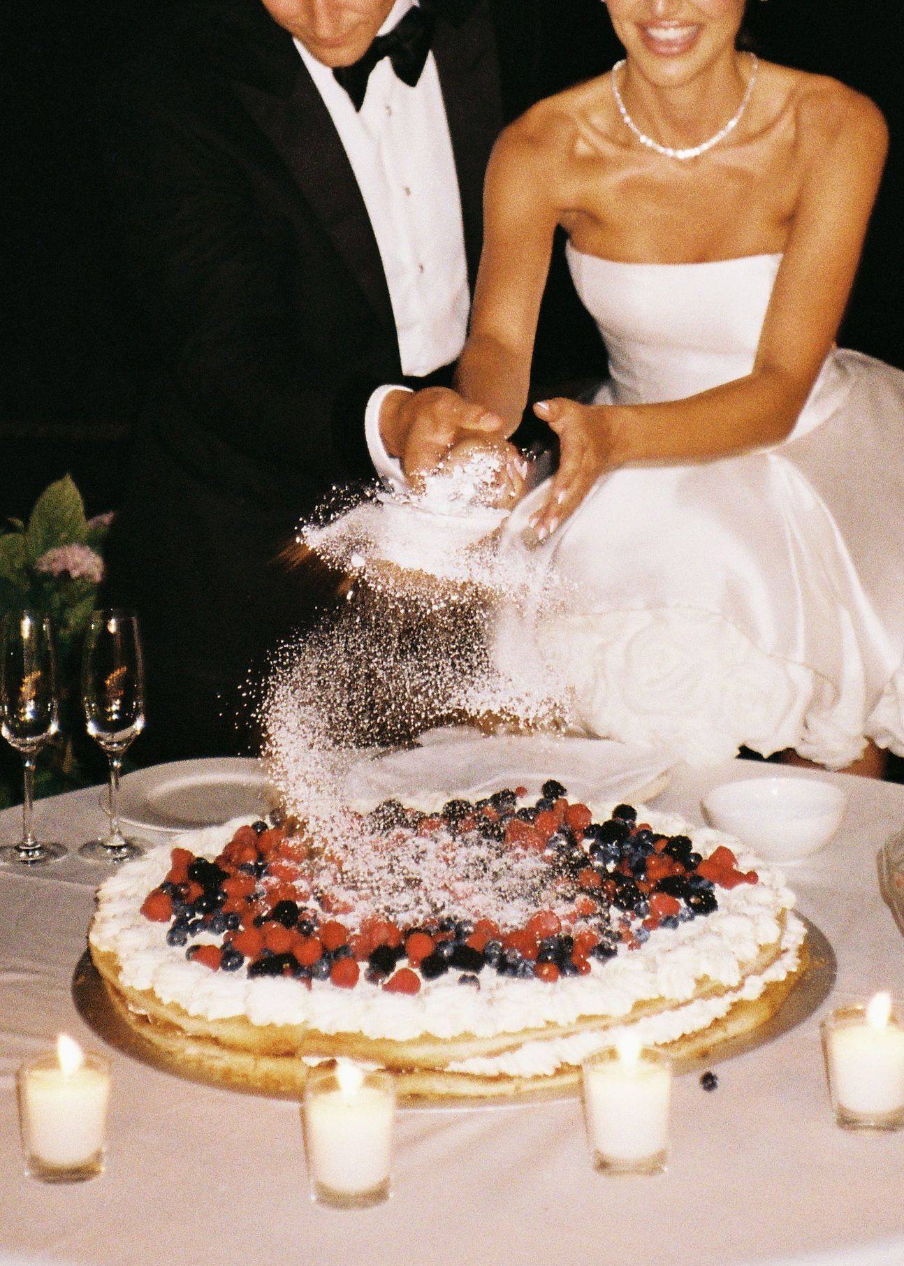 Bride and groom cutting a elegant wedding tart topped with fresh berries and powdered sugar, surrounded by candles.