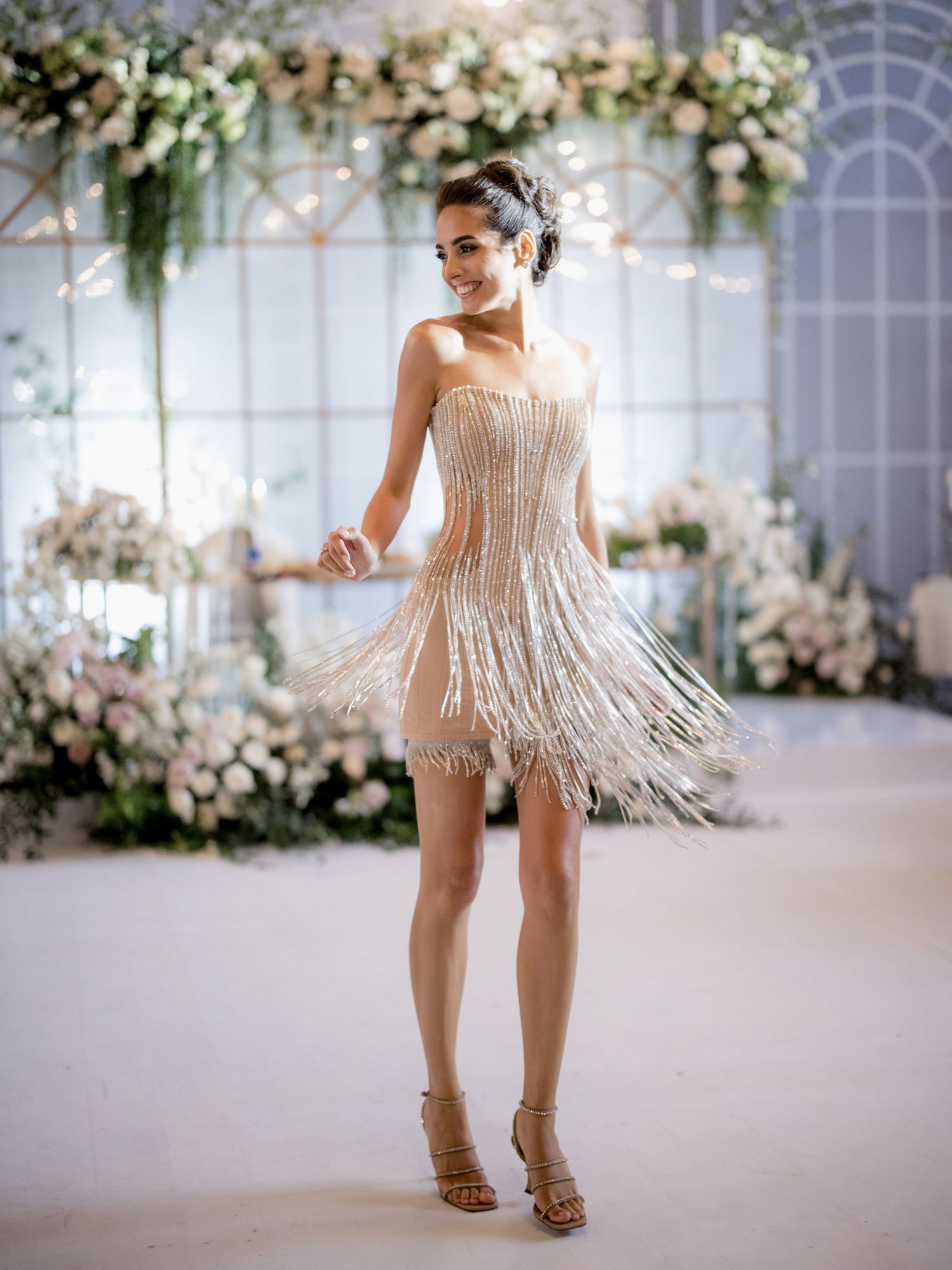Person wearing a short fringed party dress twirling in front of floral wedding arch.