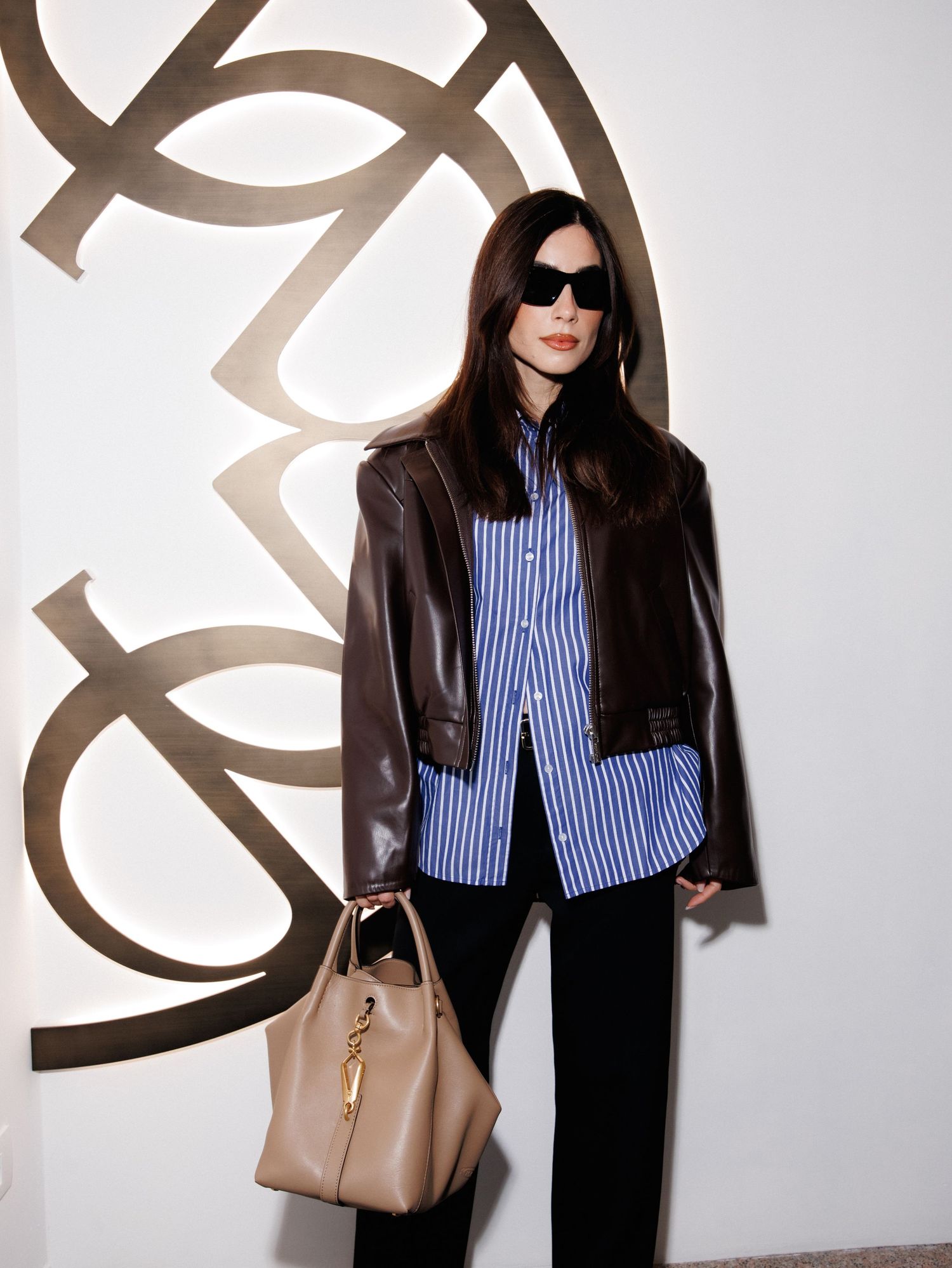Casual street style outfit featuring striped shirt, black jacket and beige handbag against white wall.