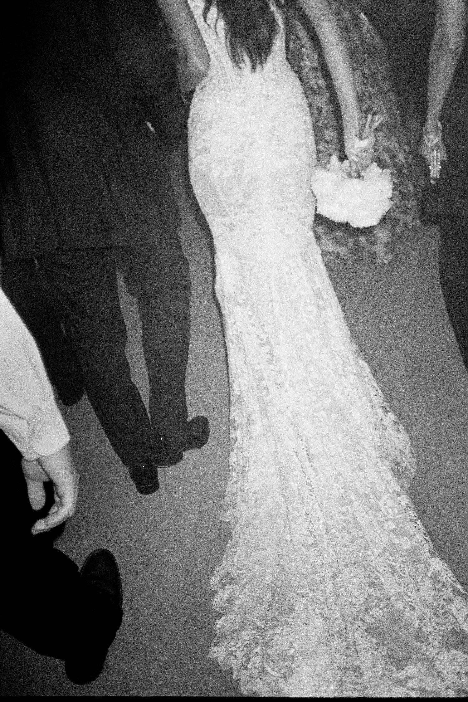 A close-up view of a white lace wedding dress train trailing on the floor during a celebration.