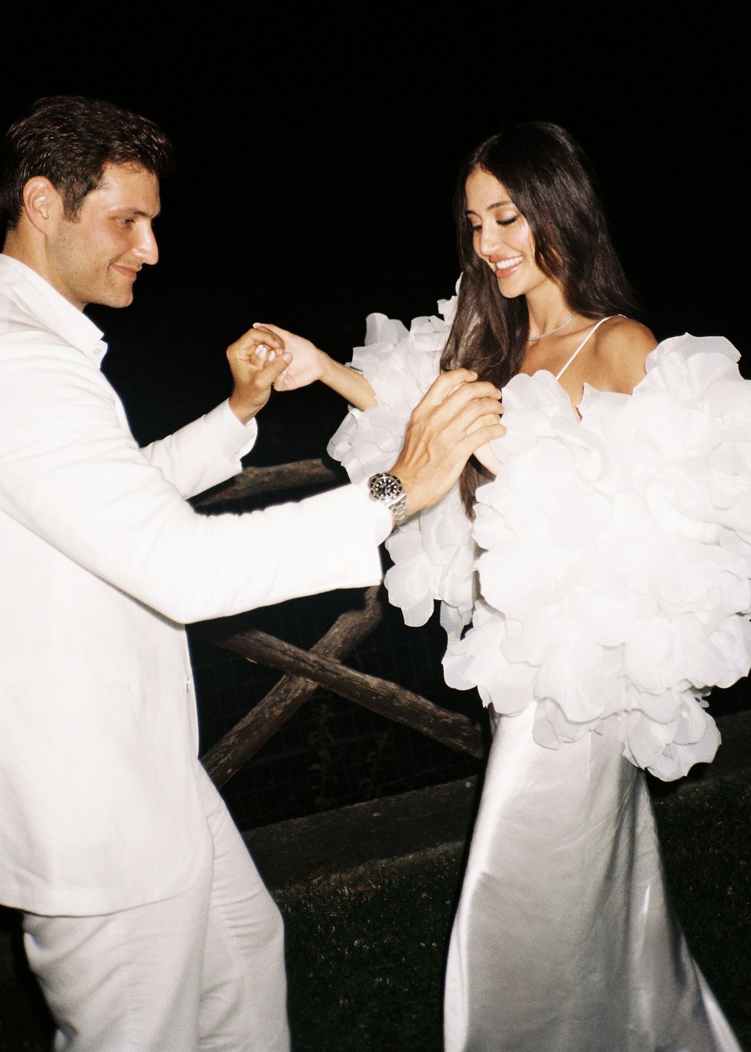 Elegant couple in white attire dancing outdoors at night, bride wearing ruffled gown, groom in white suit.