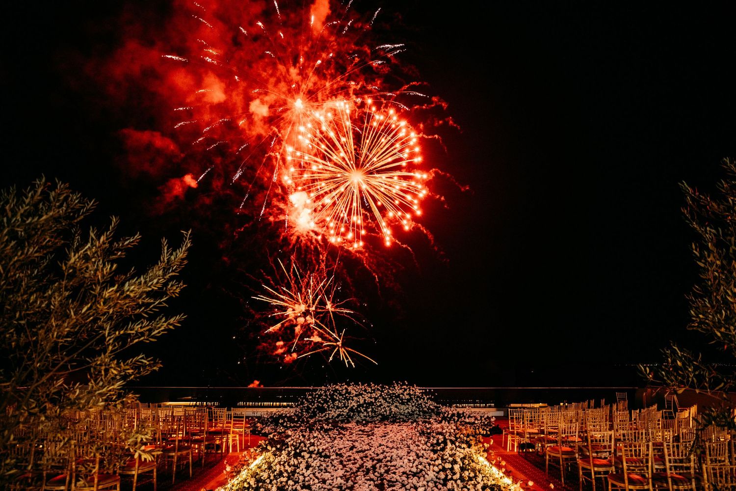 Red fireworks exploding over an outdoor wedding ceremony at night, illuminating rows of chairs and guests below.