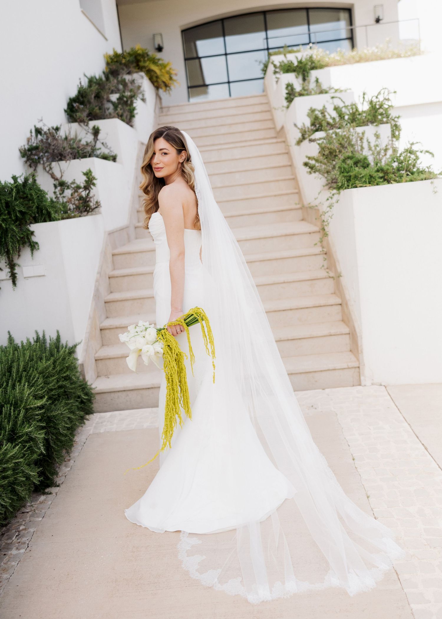 Bride in white strapless gown with long veil holds yellow floral bouquet on outdoor staircase.