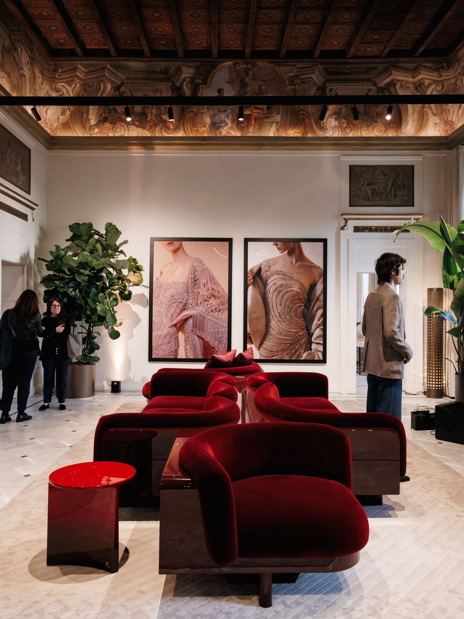 A modern art gallery space features red velvet chaise lounges arranged against white walls displaying large artwork.