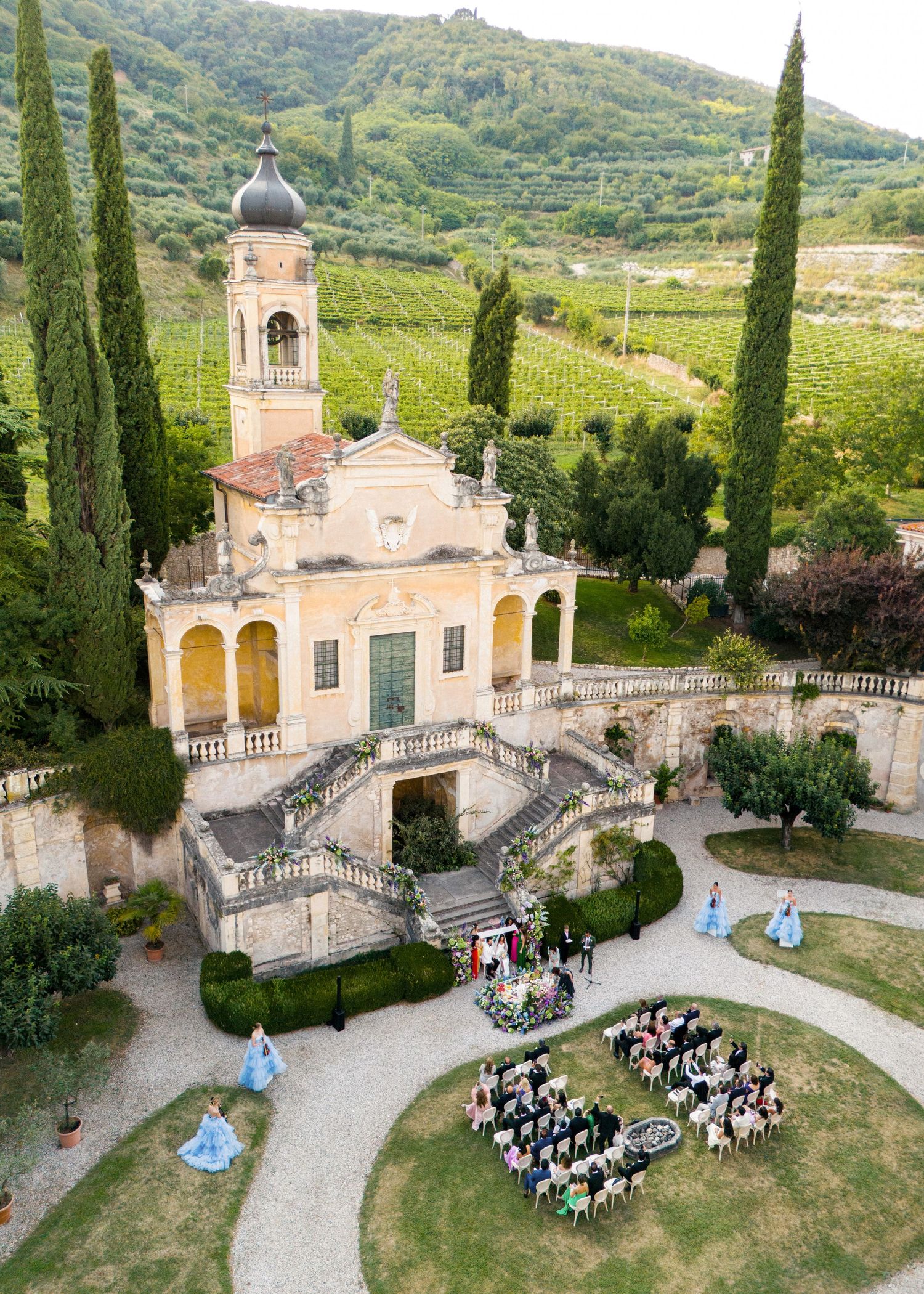Aerial view of an elegant Italian villa wedding ceremony with guests seated in a heart shape surrounded by cypress trees and vineyards.