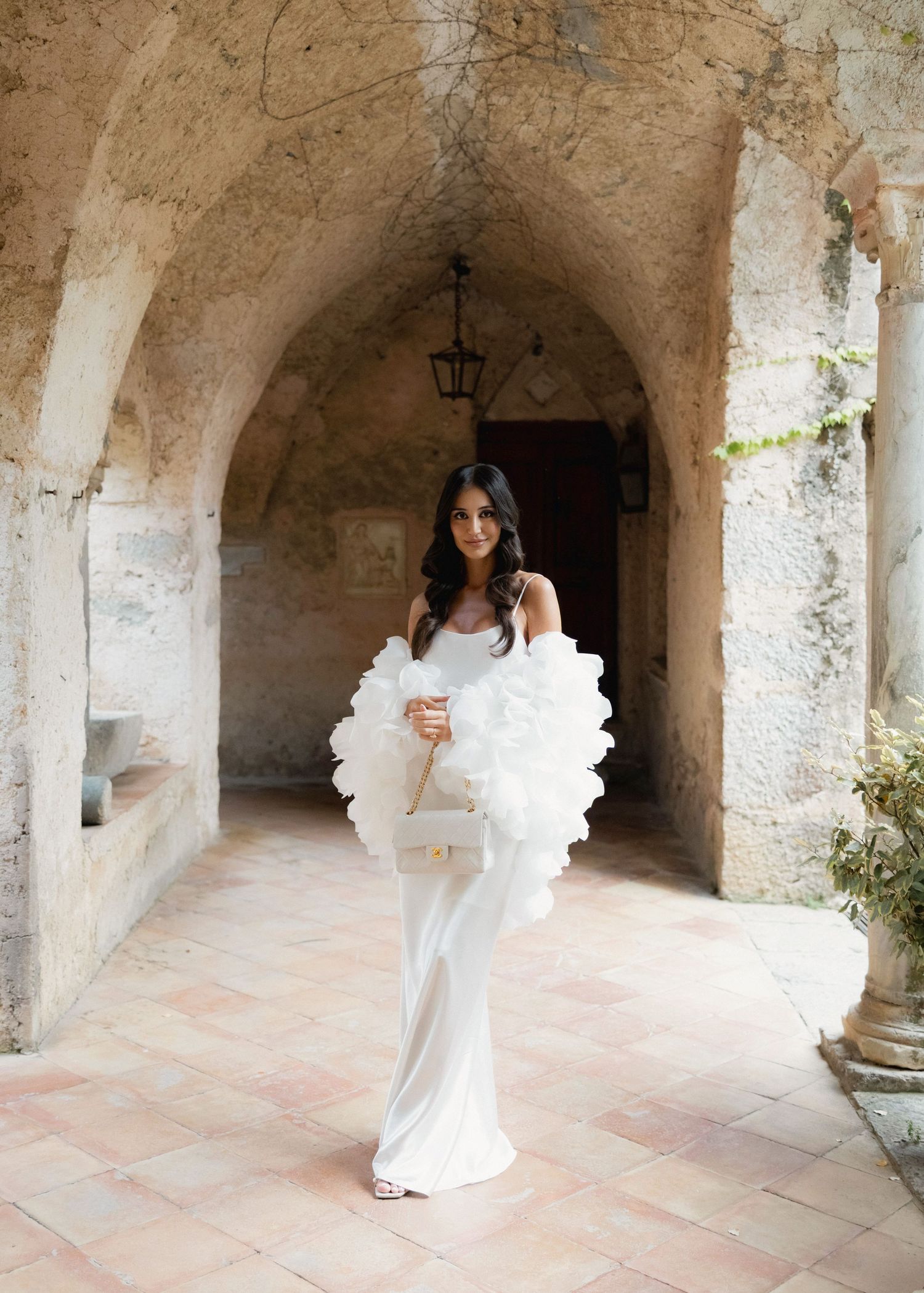 Bride in white gown holding ruffled wrap in stone archway corridor of historic venue.