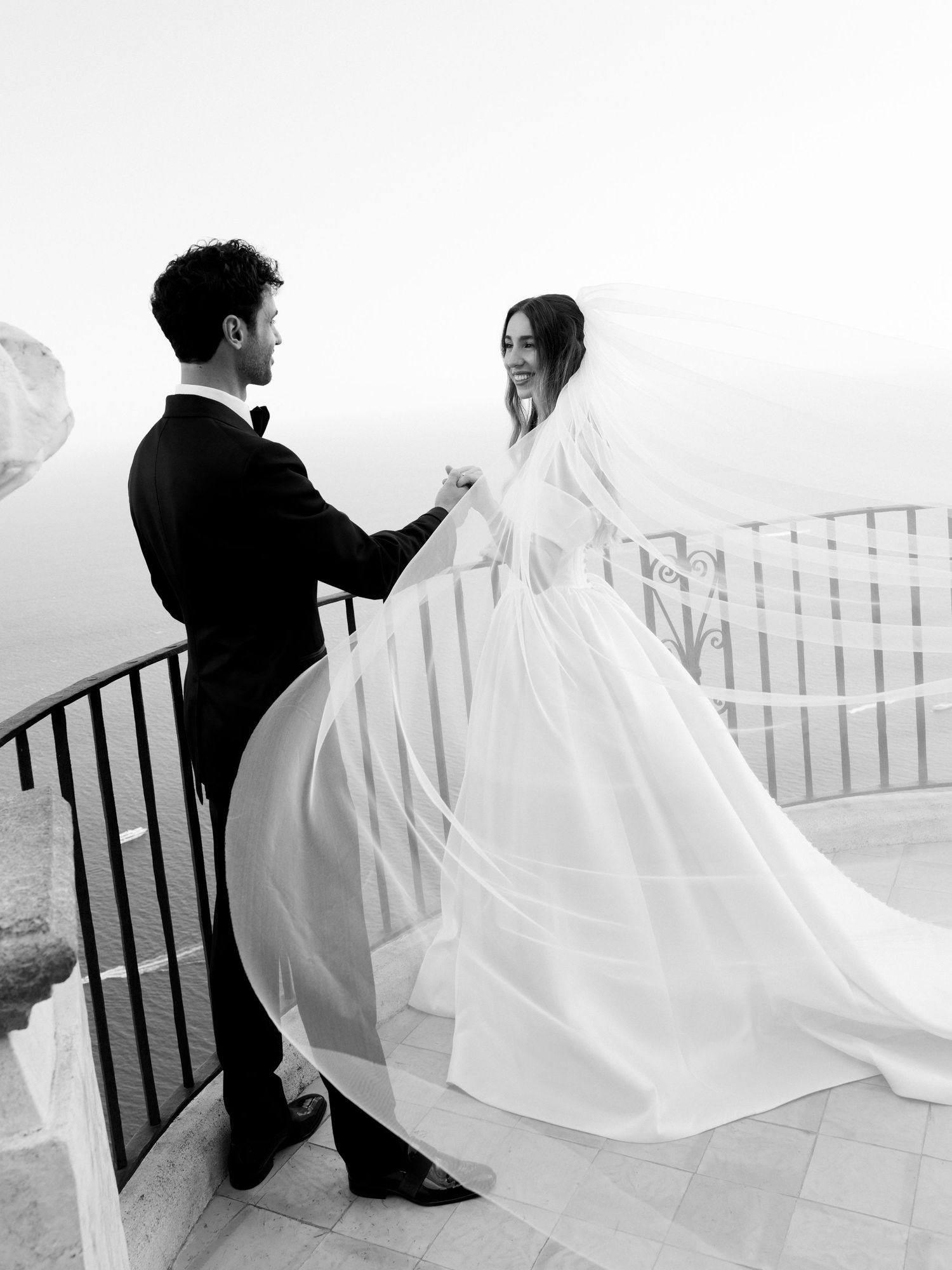 Black and white photo of a bride and groom holding hands on a scenic balcony, her veil flowing in the breeze.