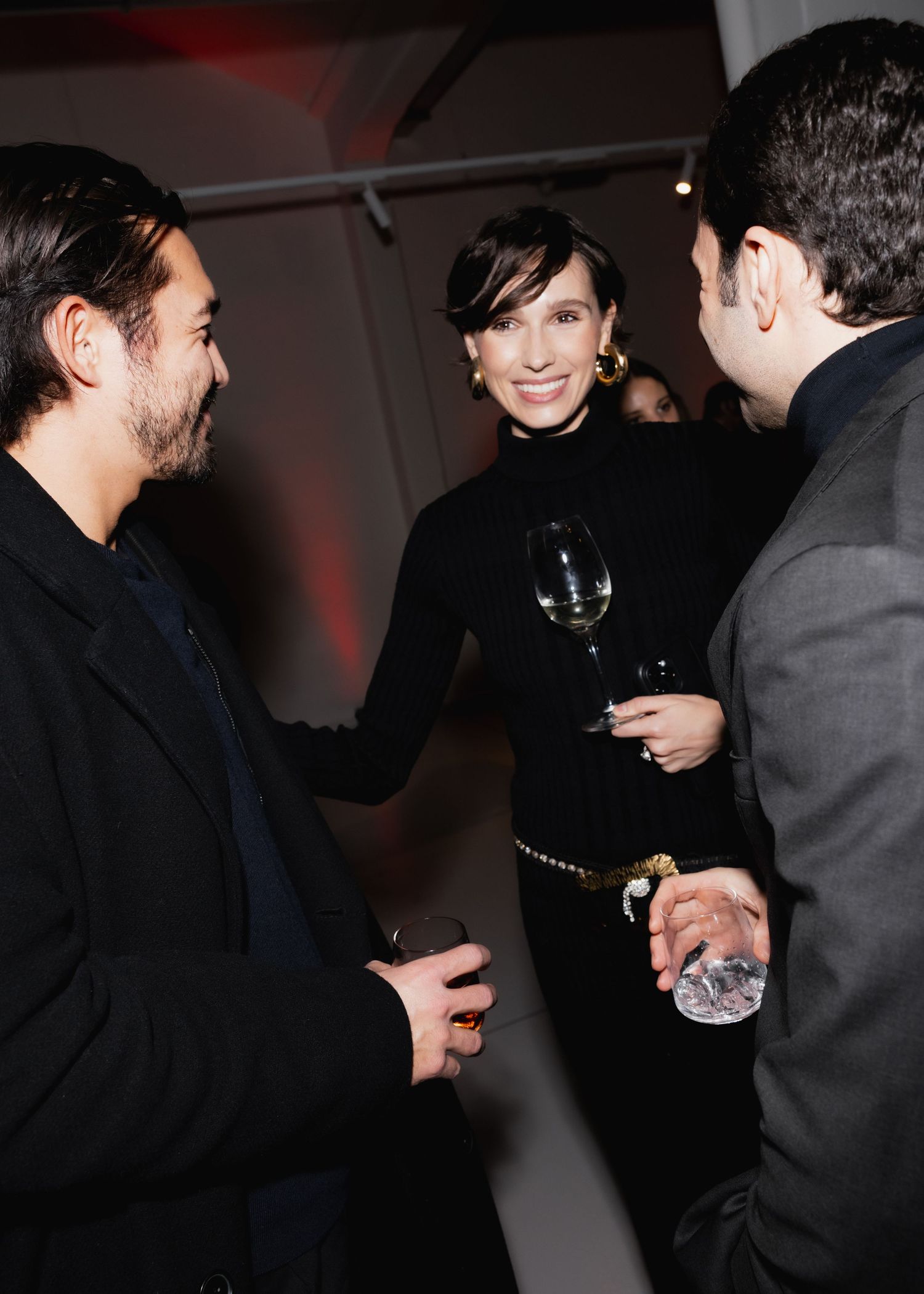 People in dark attire socialize and mingle at an indoor event with red ambient lighting.