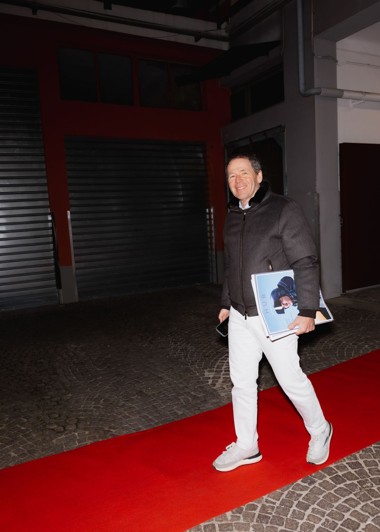 A person in casual winter wear walks along a red carpet holding papers in an industrial space.