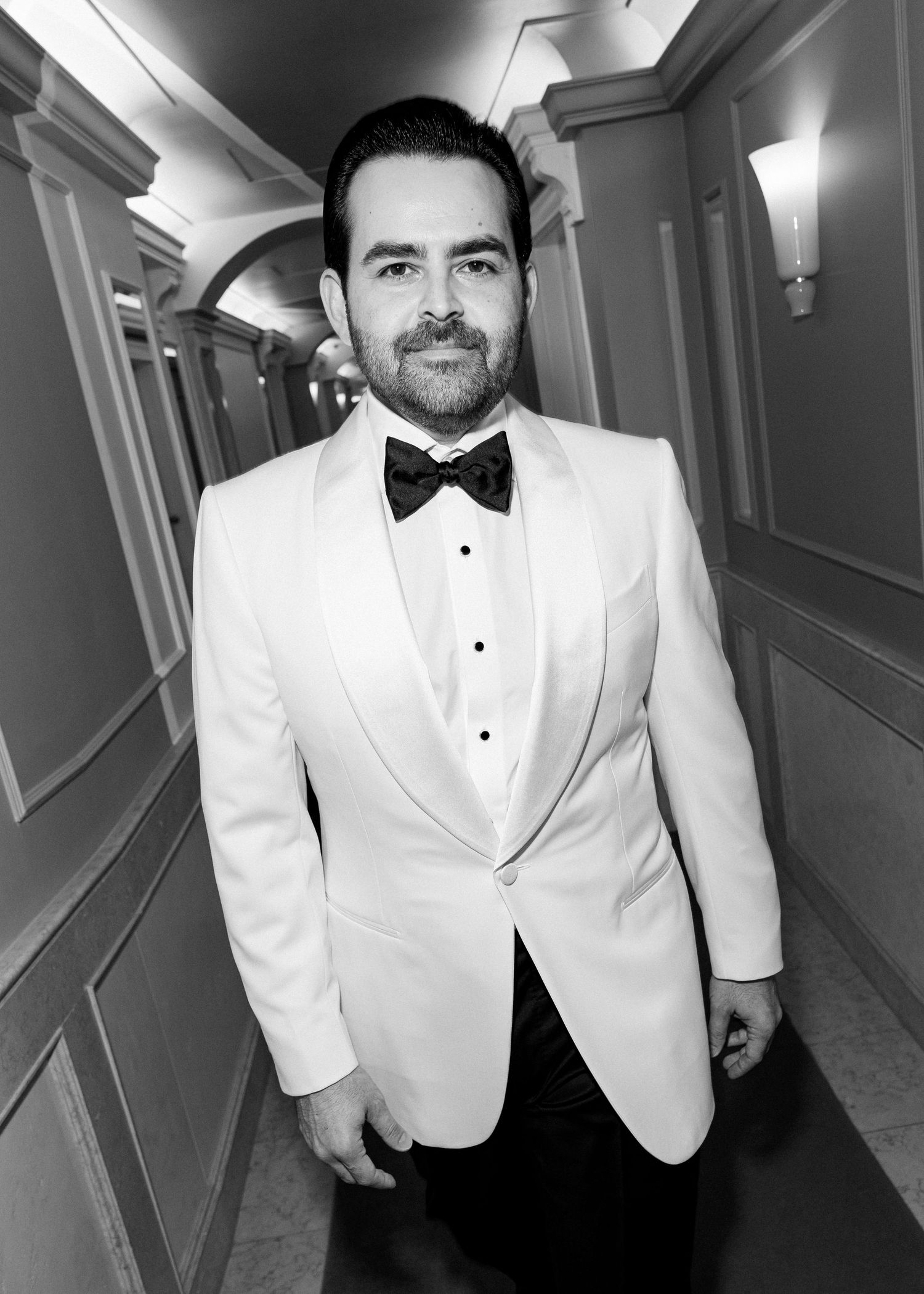 Man in white tuxedo jacket and black bow tie posing in elegant hallway, black and white formal portrait.