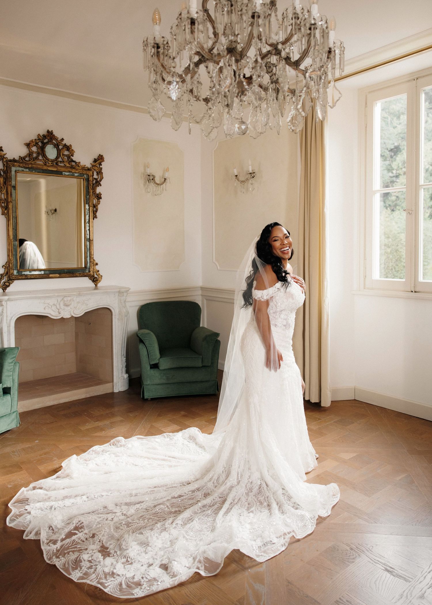 Bride in a lace wedding gown with cathedral train stands in an elegant room with chandelier and ornate mirror.