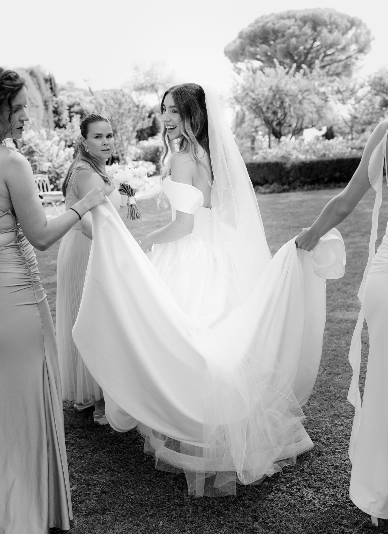 Bride in white gown and veil assisted by bridesmaids in garden, black and white wedding photo.