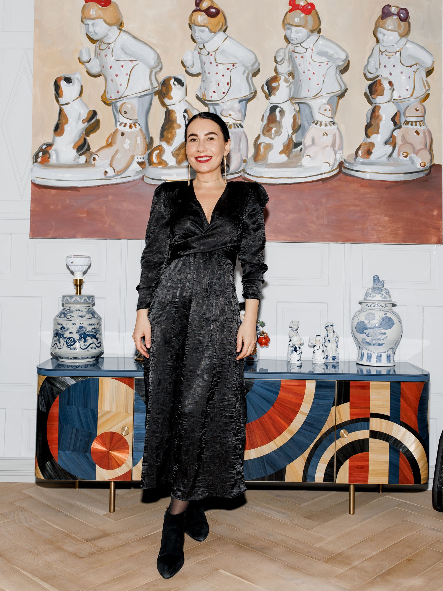 A woman in a black dress poses in front of decorative porcelain figures and art deco furniture.