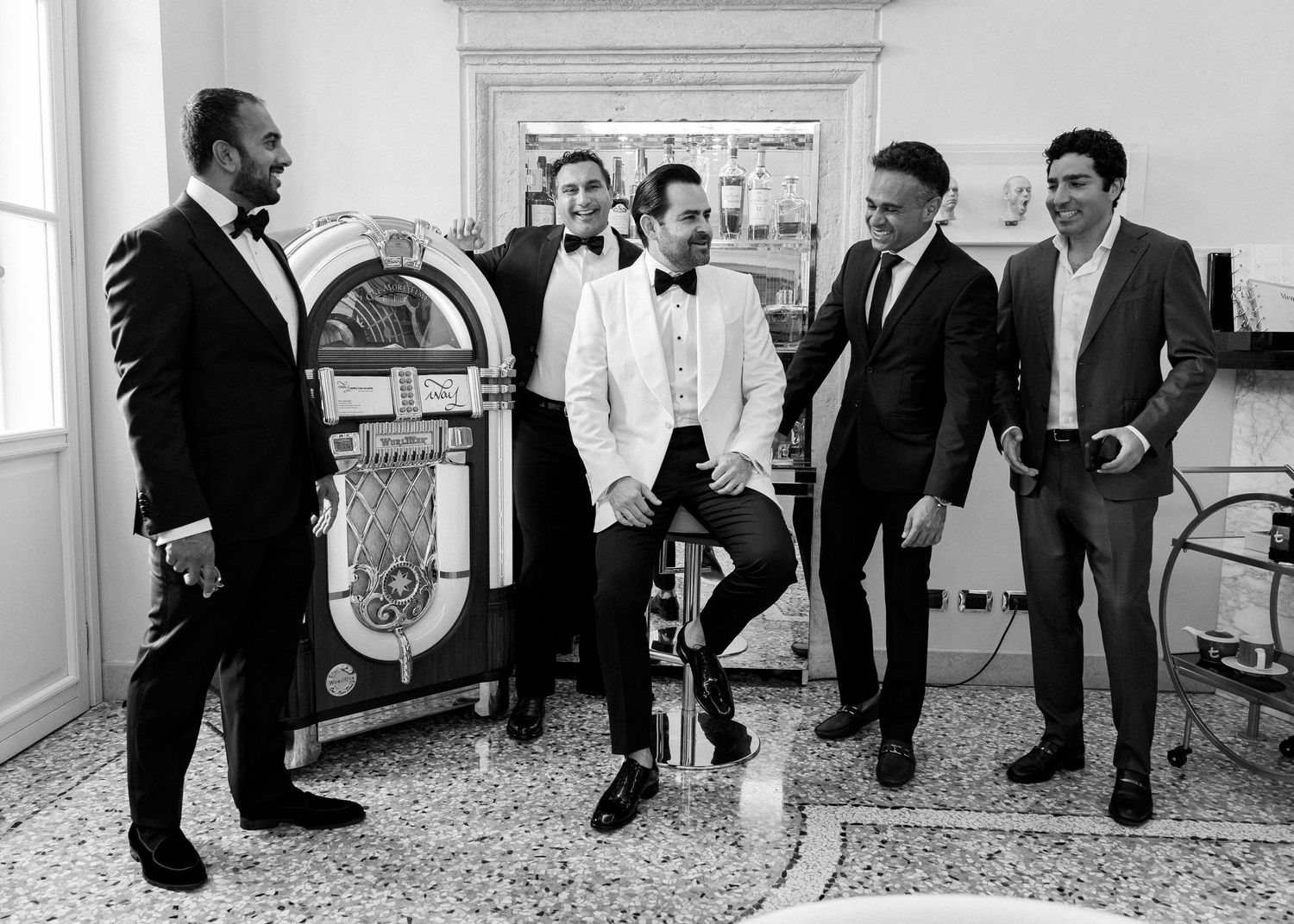 Five men in formal suits laugh together near a vintage jukebox in a black and white wedding photo.