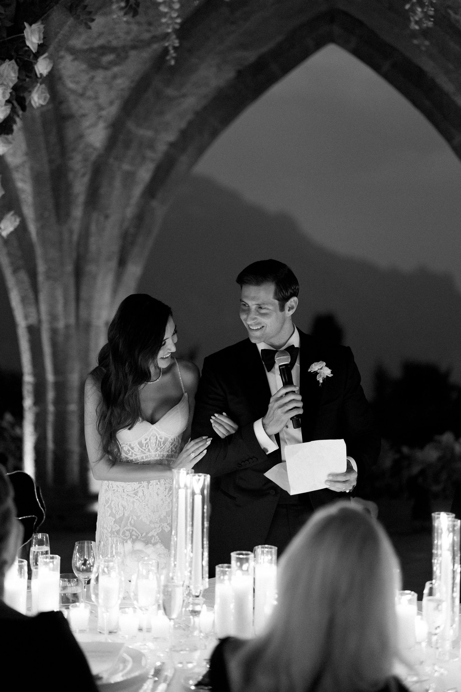 Black and white photo of a groom giving a wedding speech while bride smiles beside him, candlelit table in foreground.