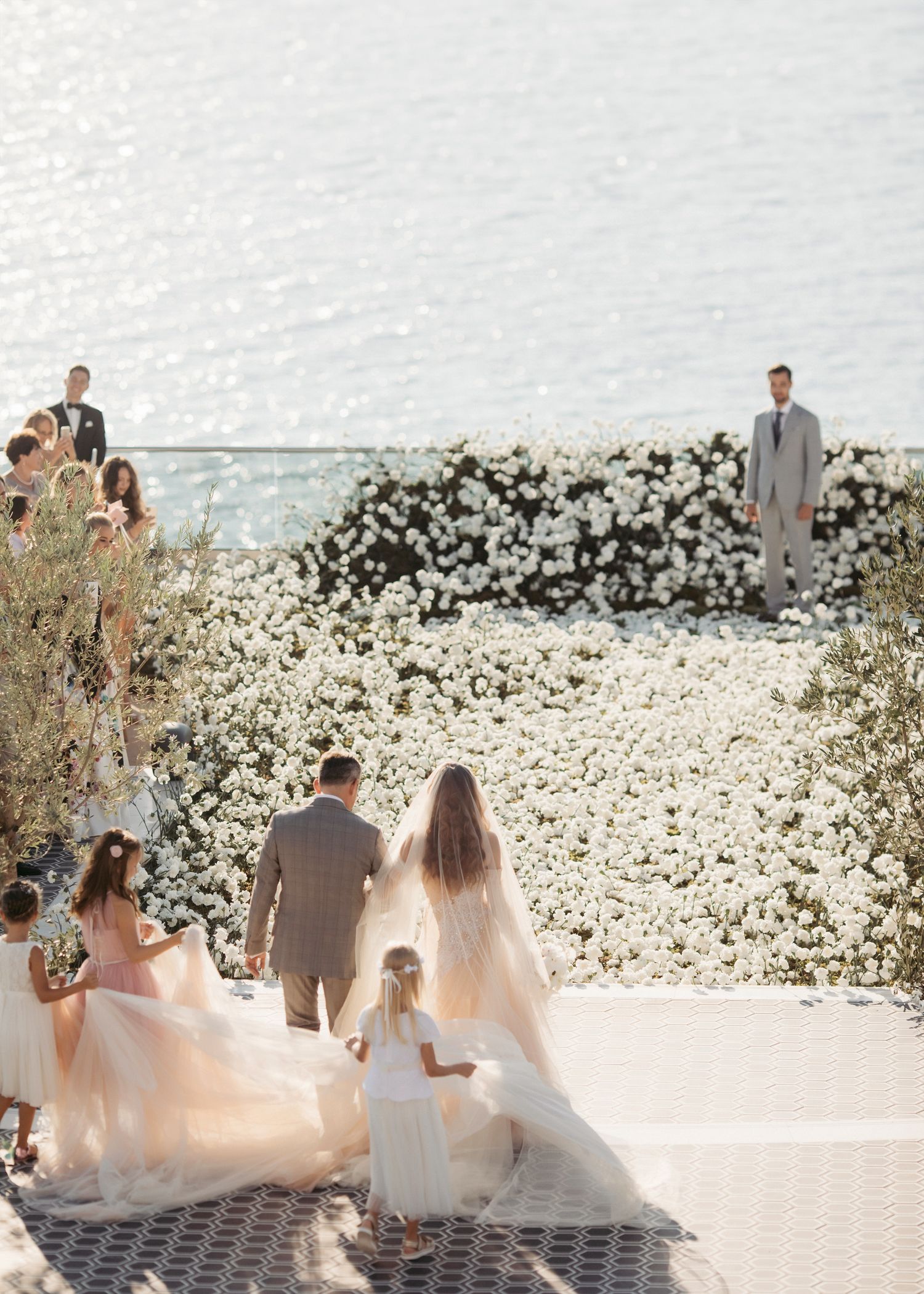 Bride and groom walk toward floral altar by the sea, accompanied by flower girls in blush dresses.