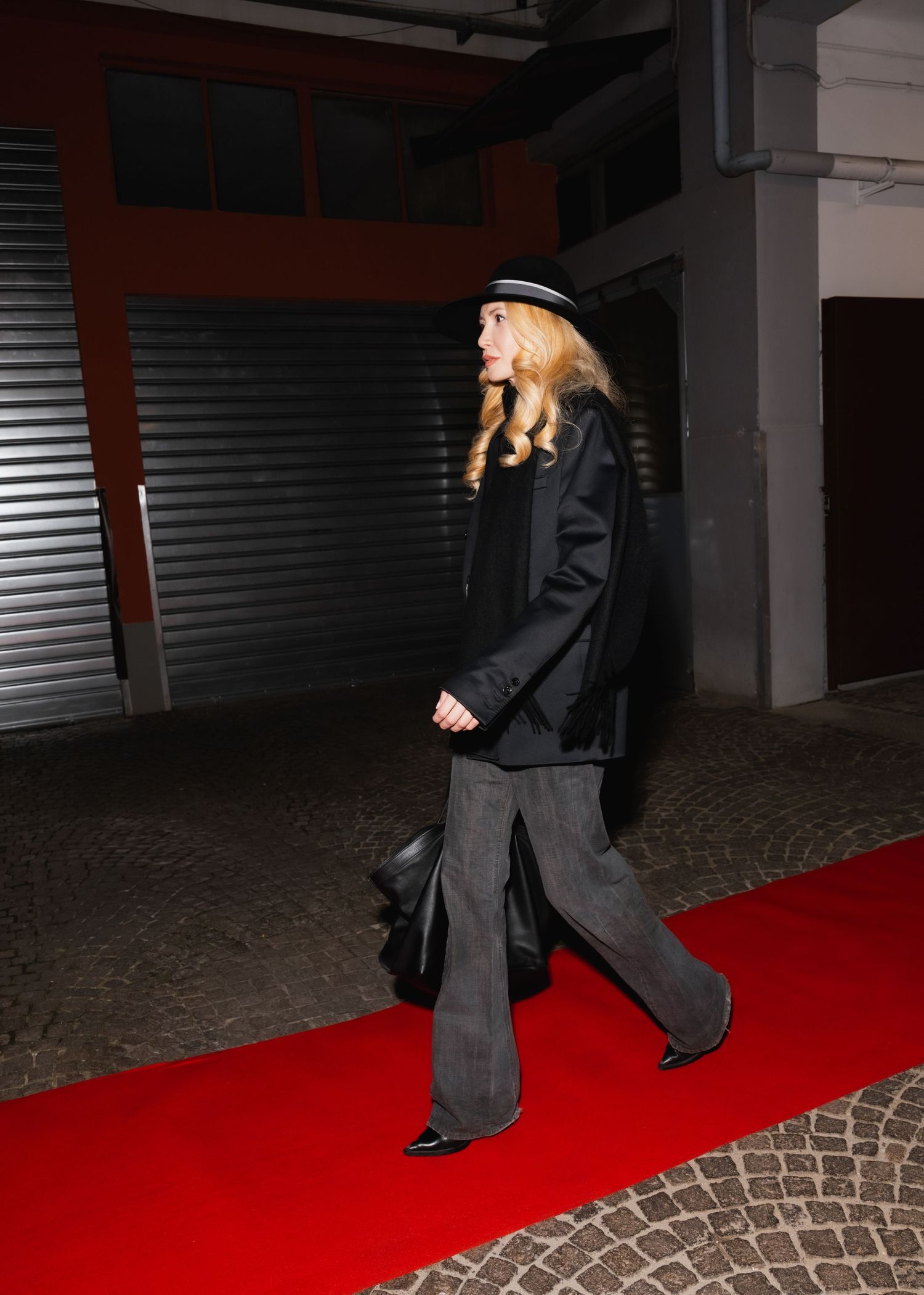 Person in dark coat walks across red carpet in dimly lit industrial space.
