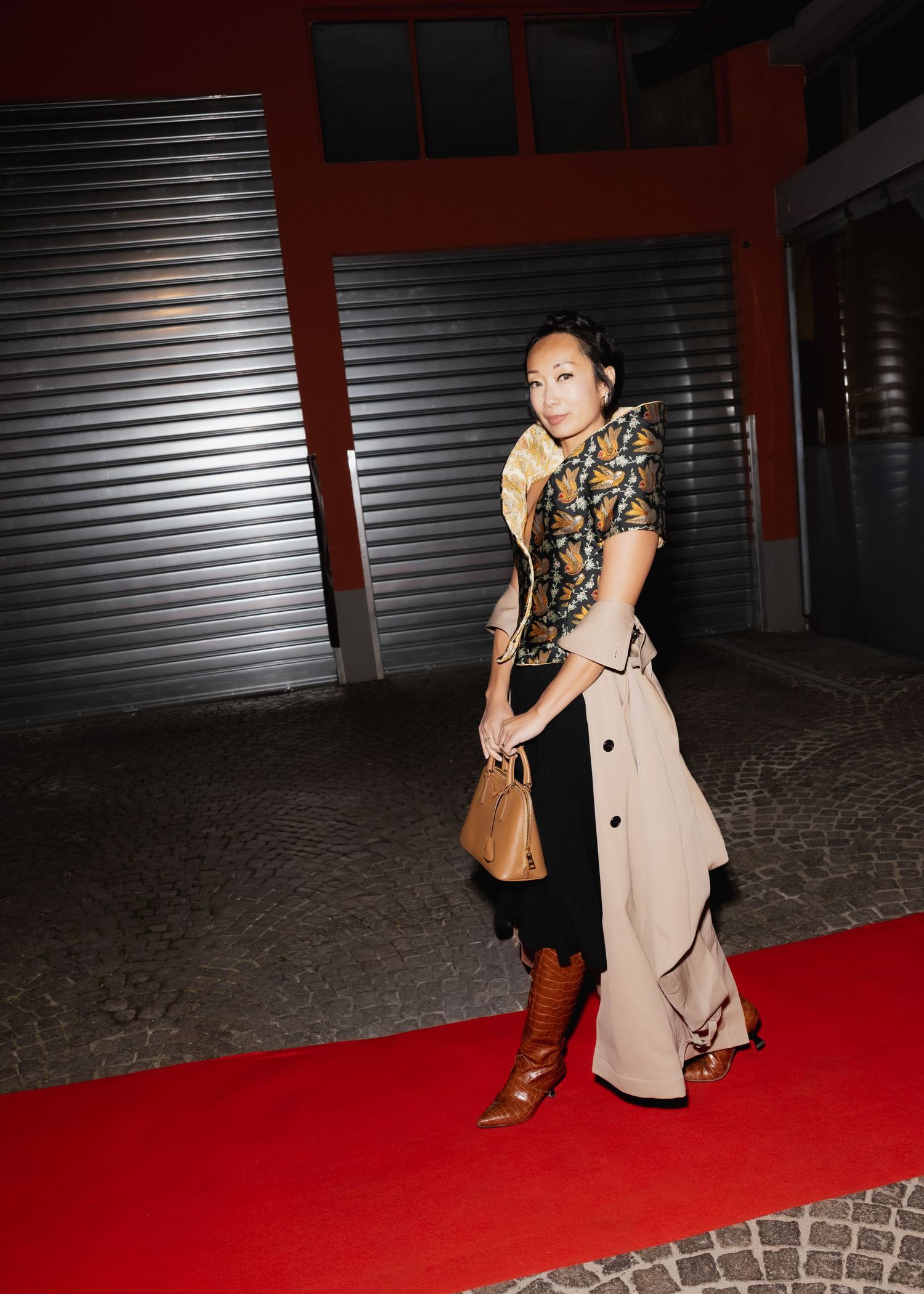 Person walking on a red carpet at night wearing a beige coat and brown leather bag.