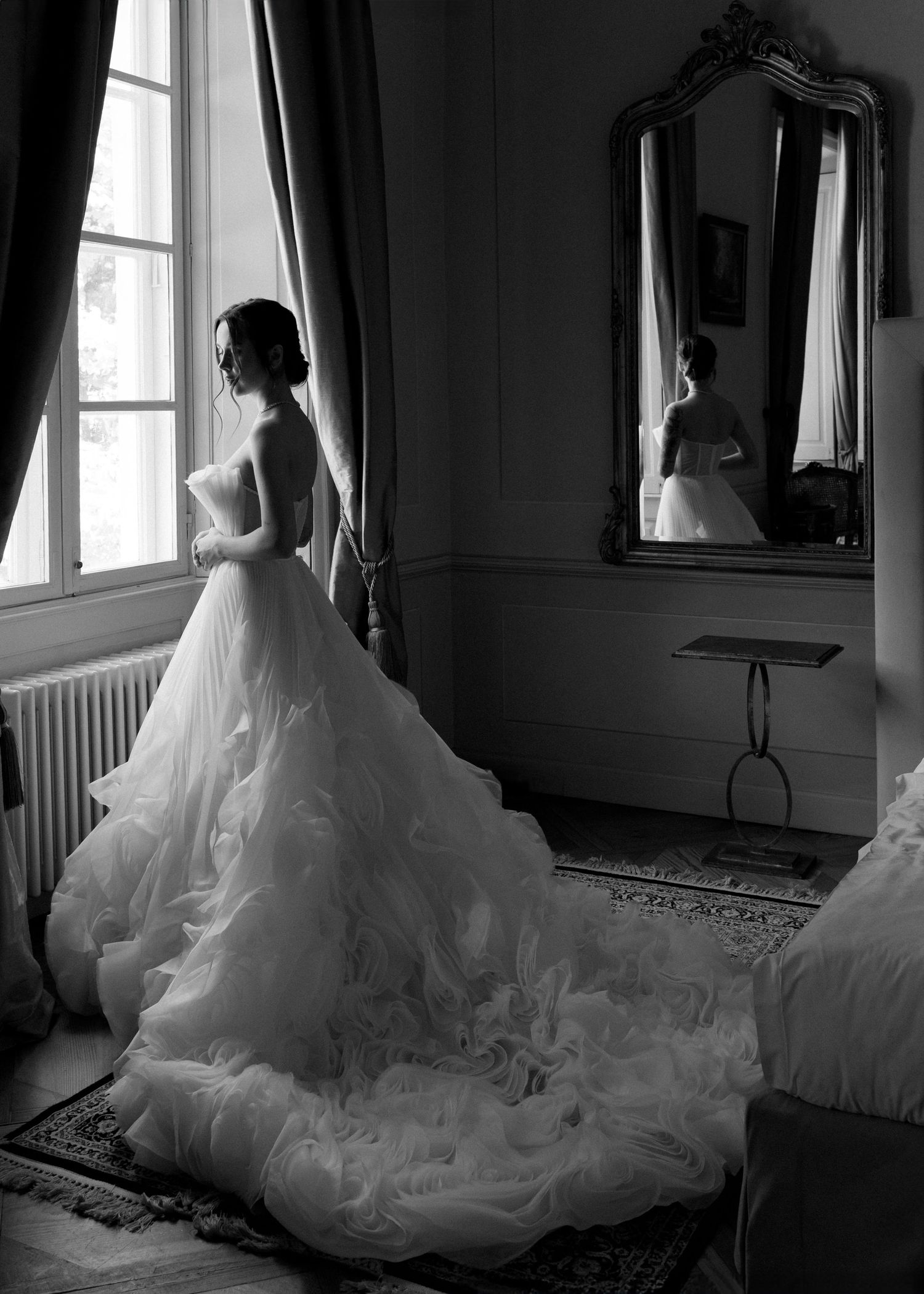 Black and white photo of a bride in a stunning ruffled ball gown with long train, standing by a window near an ornate mirror.