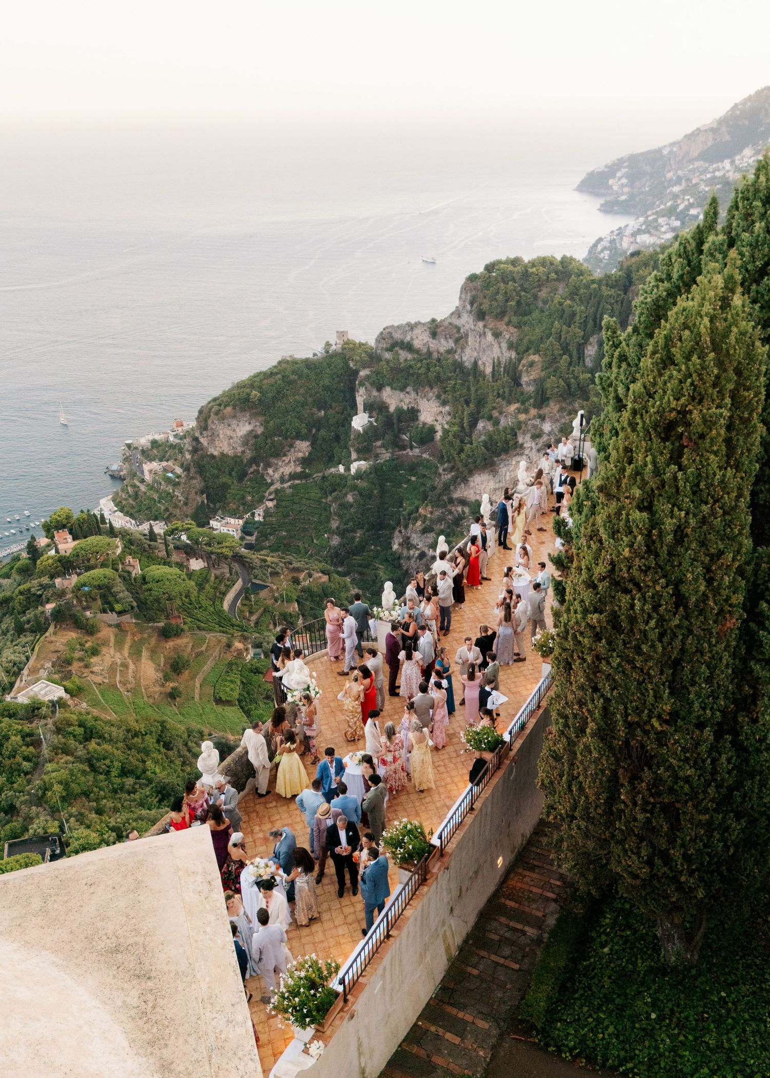 Aerial view of an elegant outdoor wedding reception on a terrace overlooking the stunning Amalfi Coast cliffs and sea.