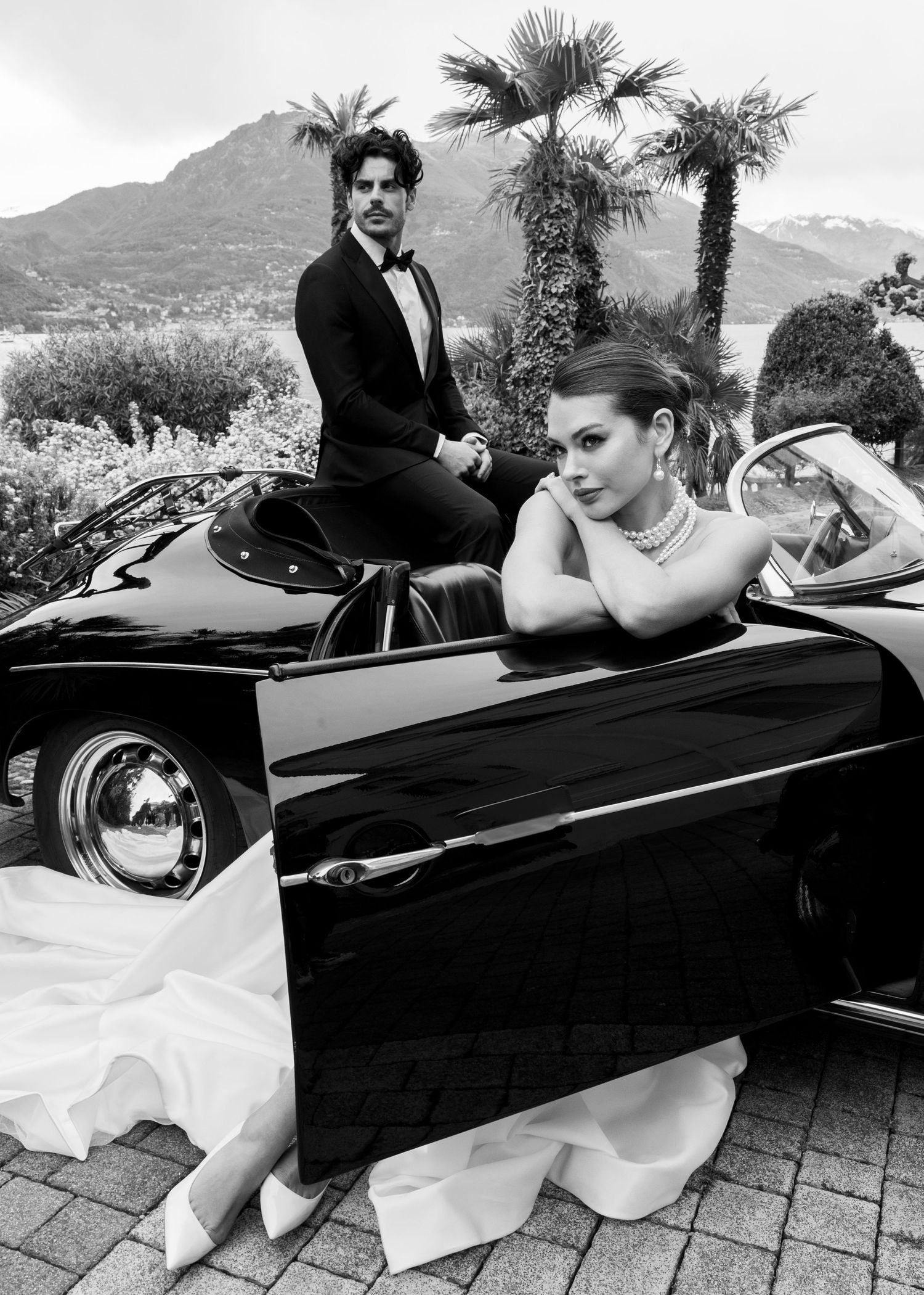 Elegant couple in formal attire posing with a classic black convertible car, black and white photo with palm trees.