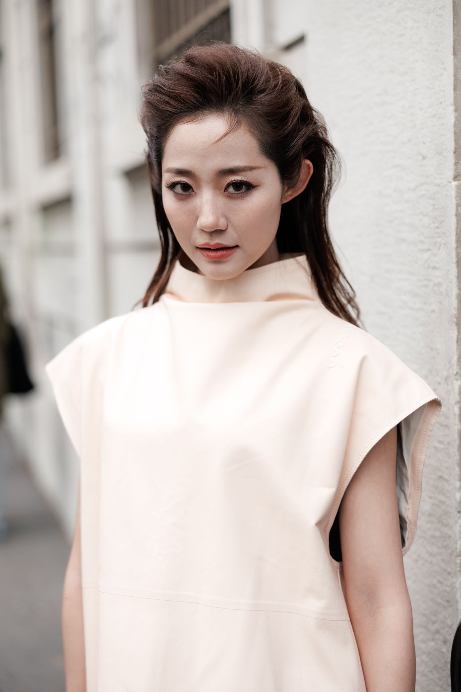 Portrait in soft lighting wearing cream-colored minimalist top against white wall.