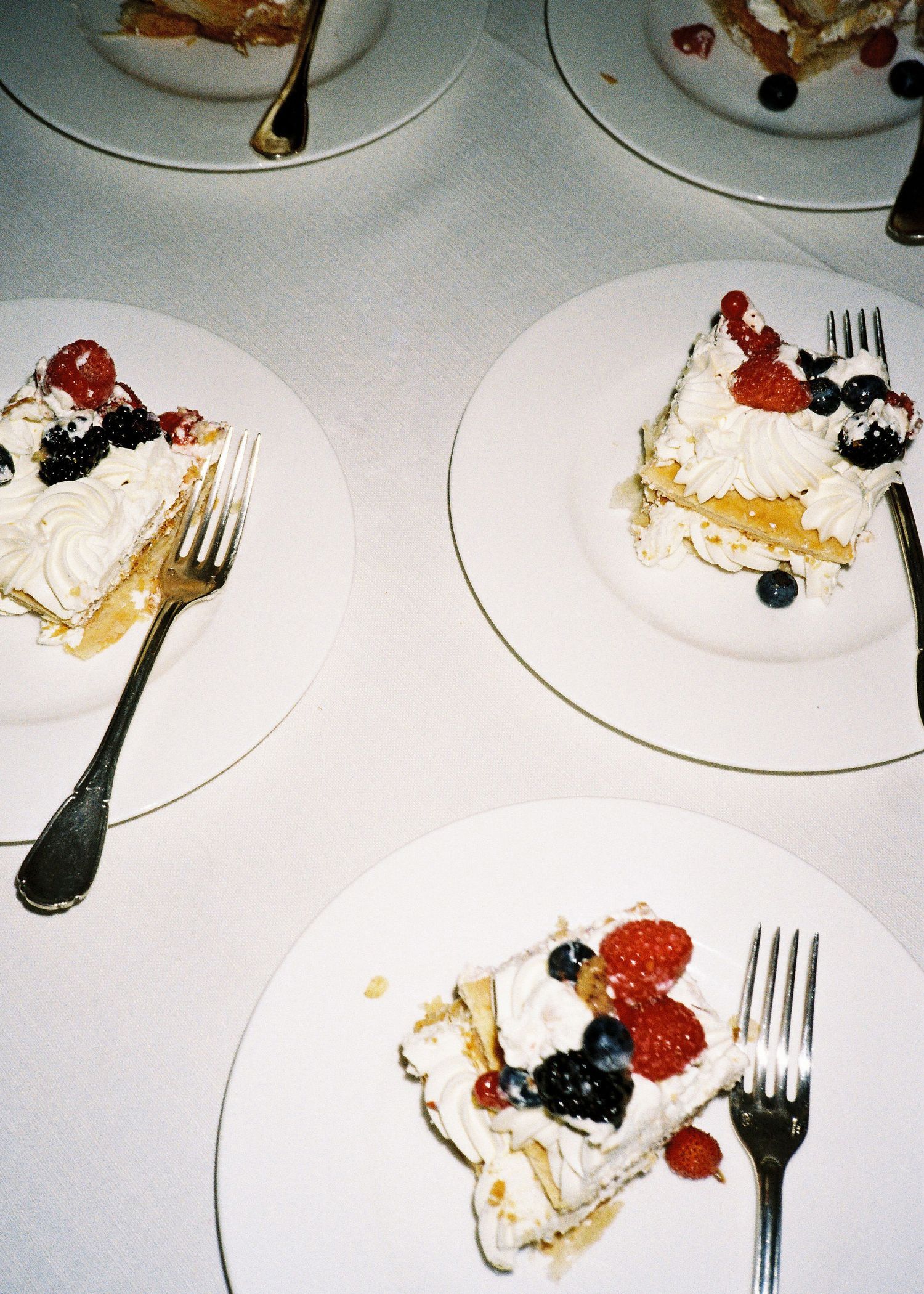 Three white plates with slices of layered cake topped with whipped cream, fresh raspberries, and blueberries on a white table.