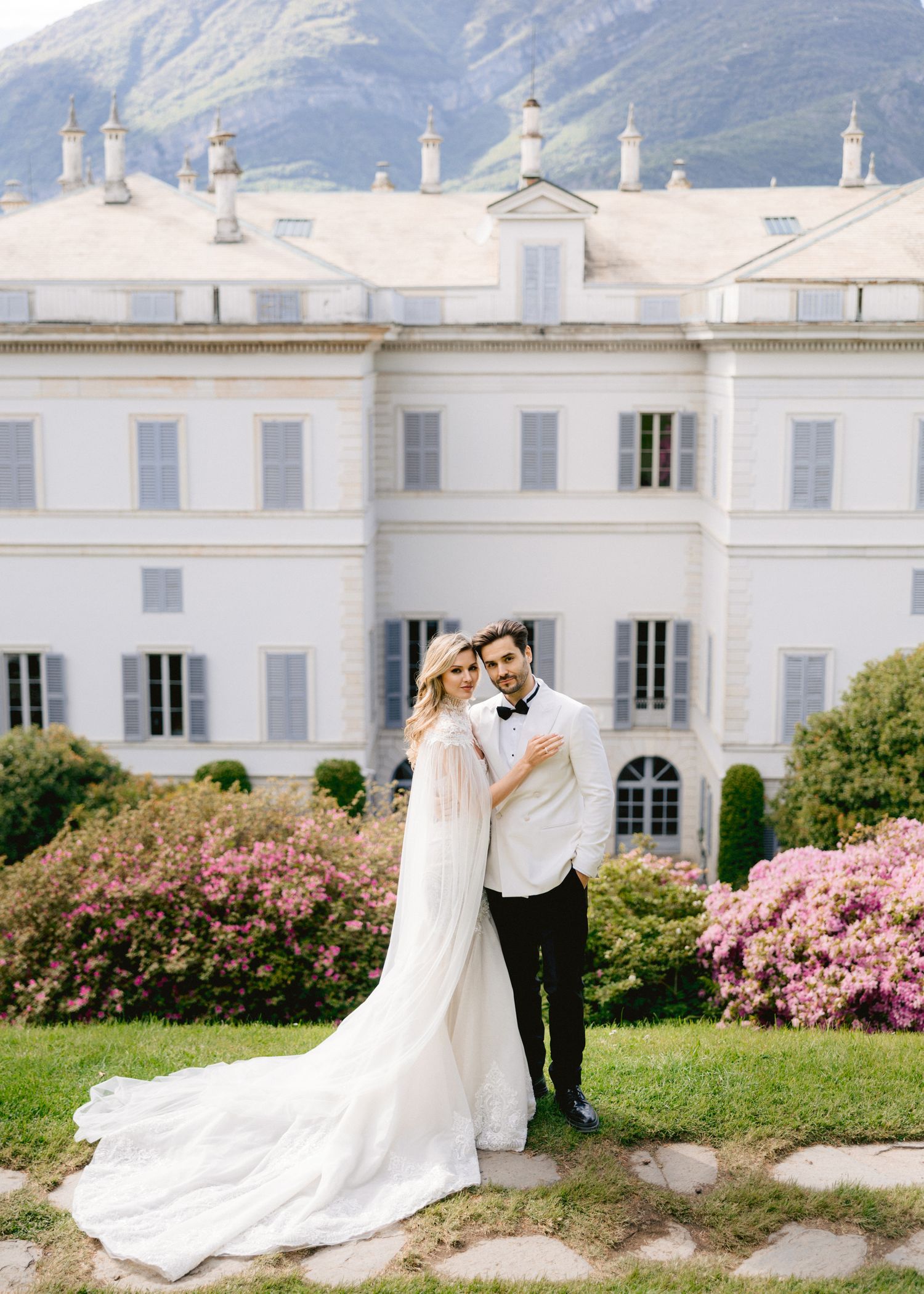 Elegant bride and groom posing in front of a grand white villa with blooming pink flowers and mountain backdrop.