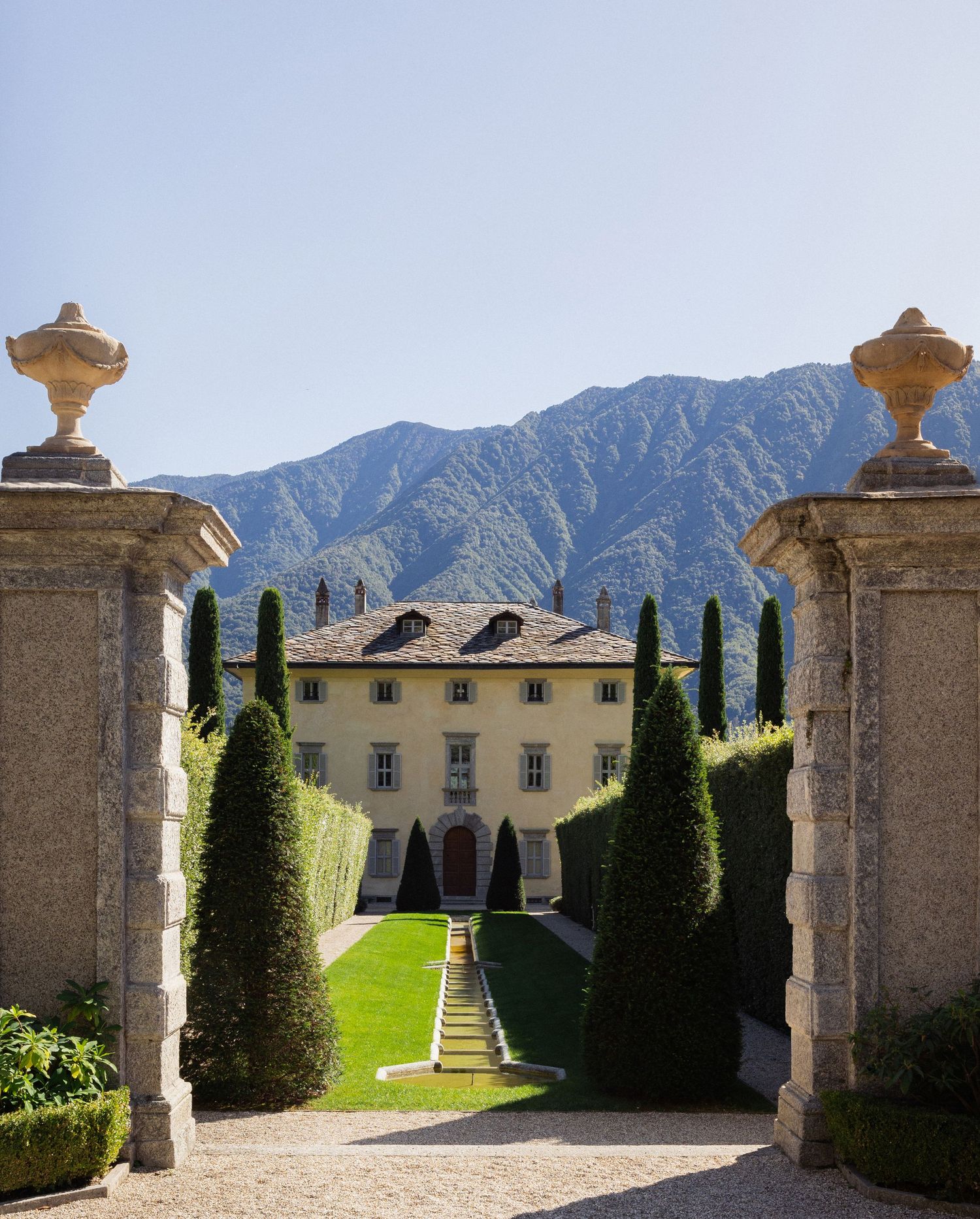 Elegant Italian villa with formal garden, cypress trees, and dramatic mountain backdrop under clear blue sky.